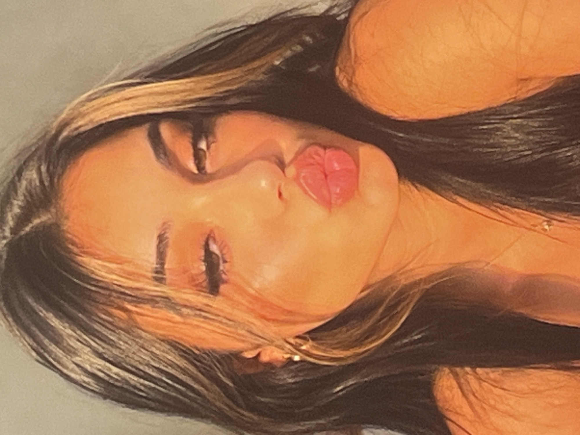 A young woman with long, dark hair featuring blonde highlights is captured in a close-up portrait. She is puckering her lips, and her eyes are partially closed. Her makeup includes dark eyeliner and mascara, and she is wearing a small gold hoop earring. The lighting in the image casts a warm, orange hue over her skin. The background is a plain, light-colored wall.
