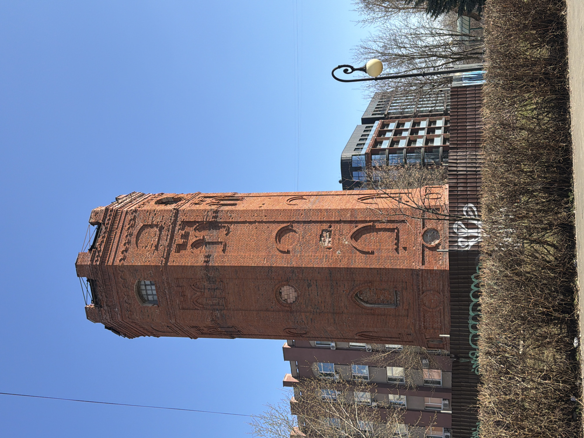 The image depicts a water tower in Izhevsk, Russia, under a clear blue sky. The tower is made of red brick and features arched architectural details. Buildings are visible in the background on both sides of the tower, along with trees and a streetlight. A trimmed hedge runs along the side of the image.