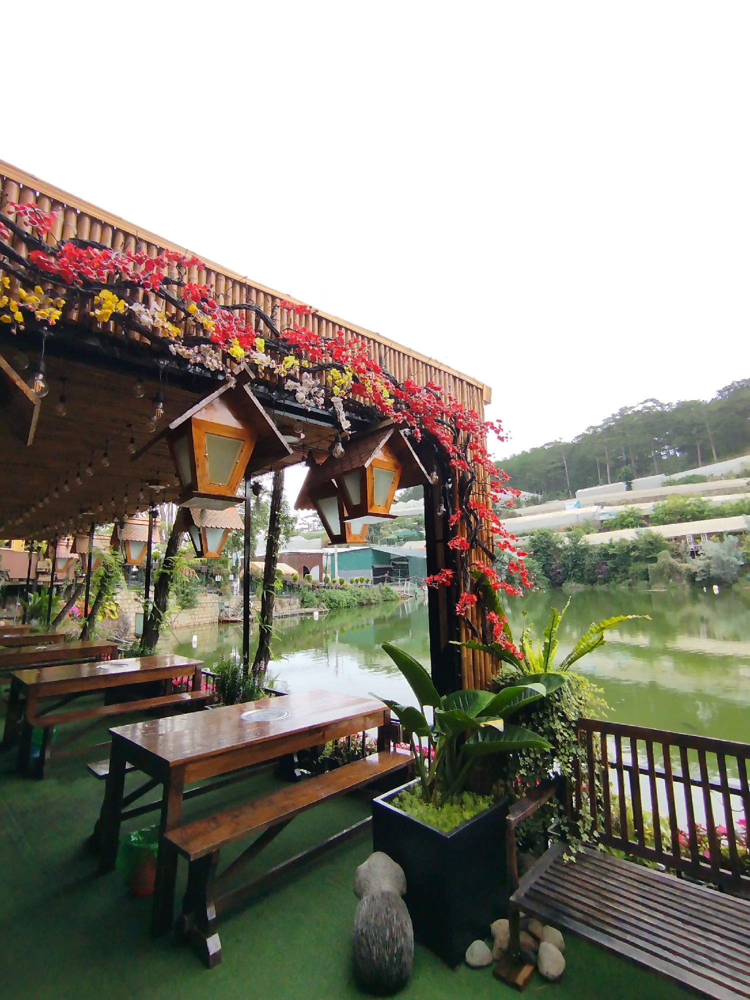 The image shows an outdoor seating area with wooden tables and benches, covered by a wooden roof adorned with colorful flowers. Traditional-style lanterns hang from the roof. A lake is visible in the background, reflecting the greenery. The area is decorated with potted plants and a wooden railing separates the seating from the water. A forested hillside is in the far background. The image appears to be taken at a restaurant or similar establishment in a lakeside setting, potentially in Da Lat, Vietnam. The seating appears to be vacant.