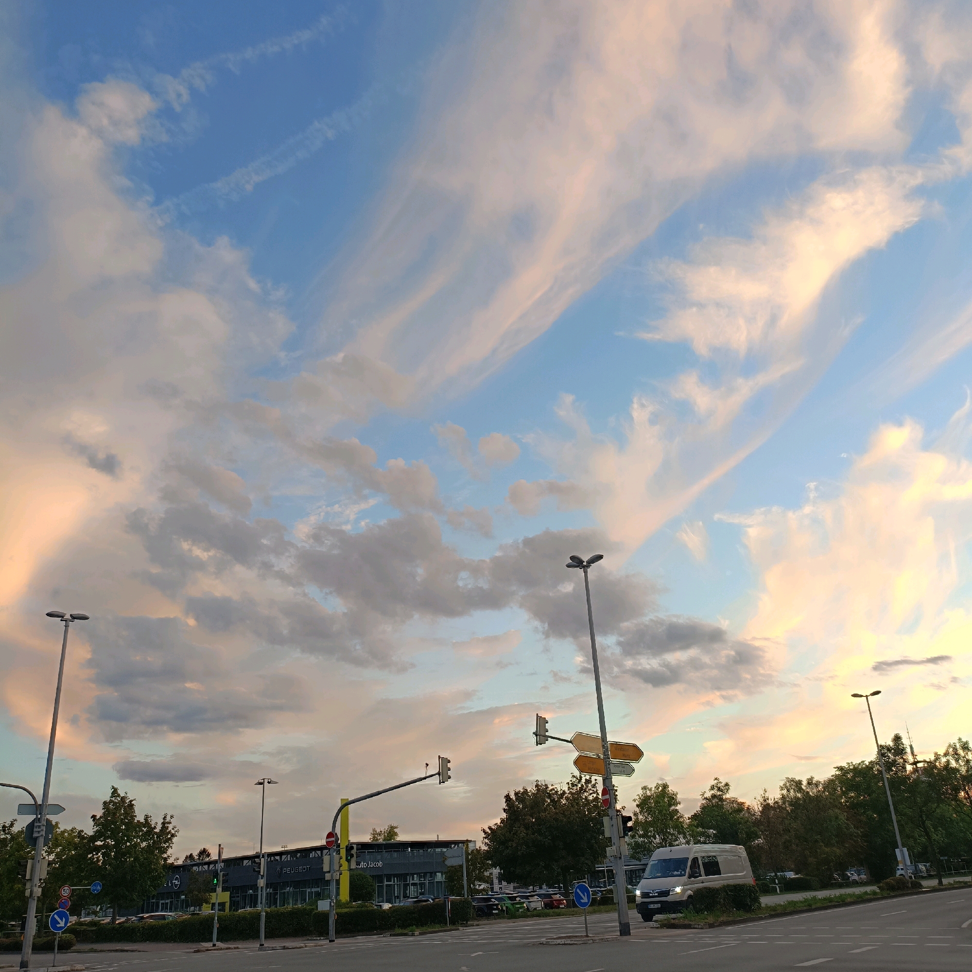The image depicts a street scene in Rüsselsheim am Main, Germany, featuring a twilight sky with wispy, pink-tinged clouds. In the foreground, there is a road intersection with traffic lights, streetlights, and traffic signs. A white van is visible driving on the right side of the road. Behind the road, a modern building with a sign that reads "PEUGEOT" and "auto Jacob" is present, suggesting it's a car dealership. Several trees and a neatly trimmed hedge line the front of the building. The overall impression is of a quiet evening in a commercial area of the city.