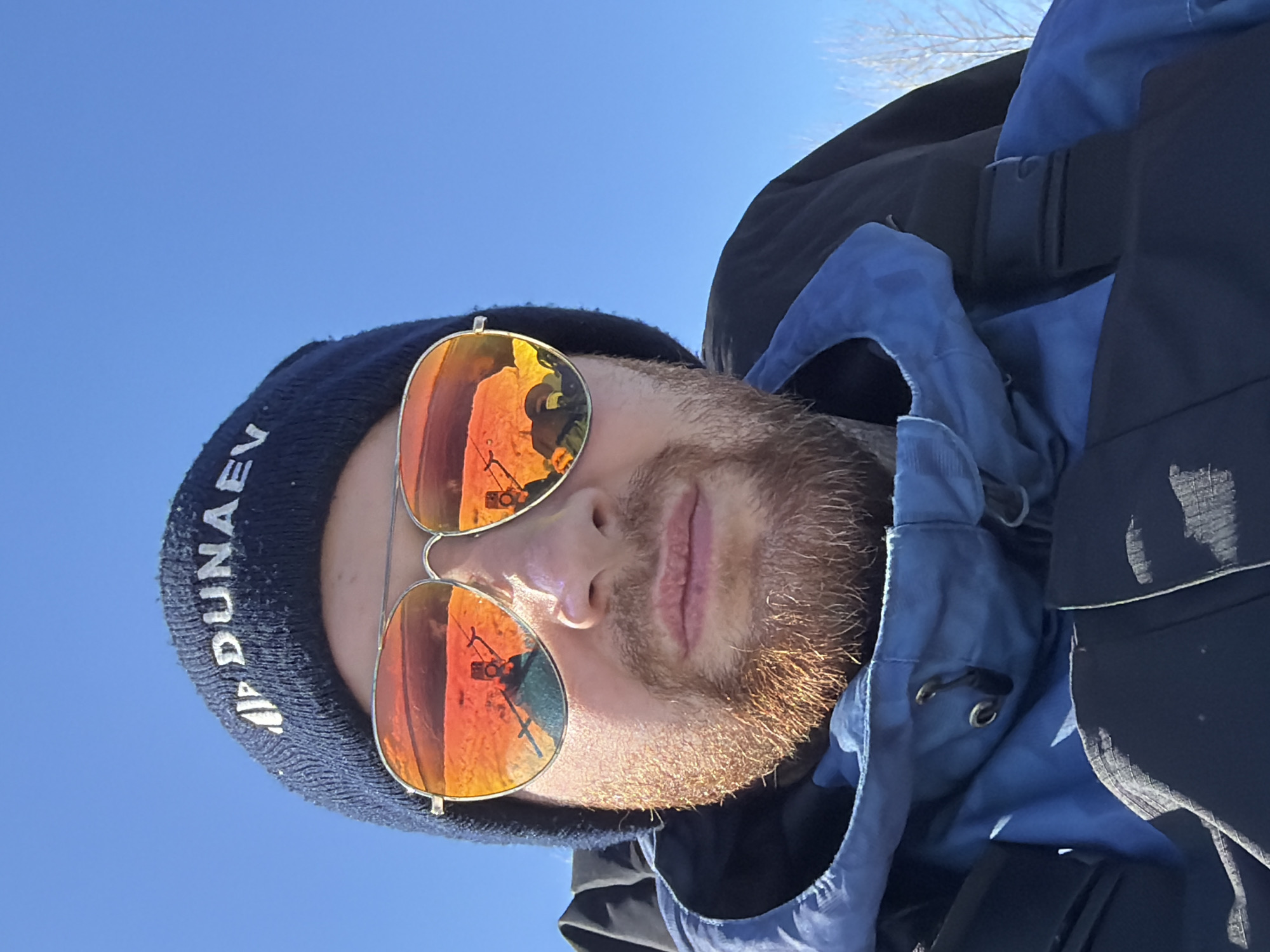 A man is taking a selfie outdoors. He is wearing a dark blue beanie with white text that includes the word "DUNAEV", aviator sunglasses with orange mirrored lenses, and a blue and black jacket. His beard is reddish-brown. The reflection in his sunglasses shows a Ferris wheel, indicating an amusement park, possibly in Chaikovsky, Russia, given the text on his beanie. The sky is clear and blue.