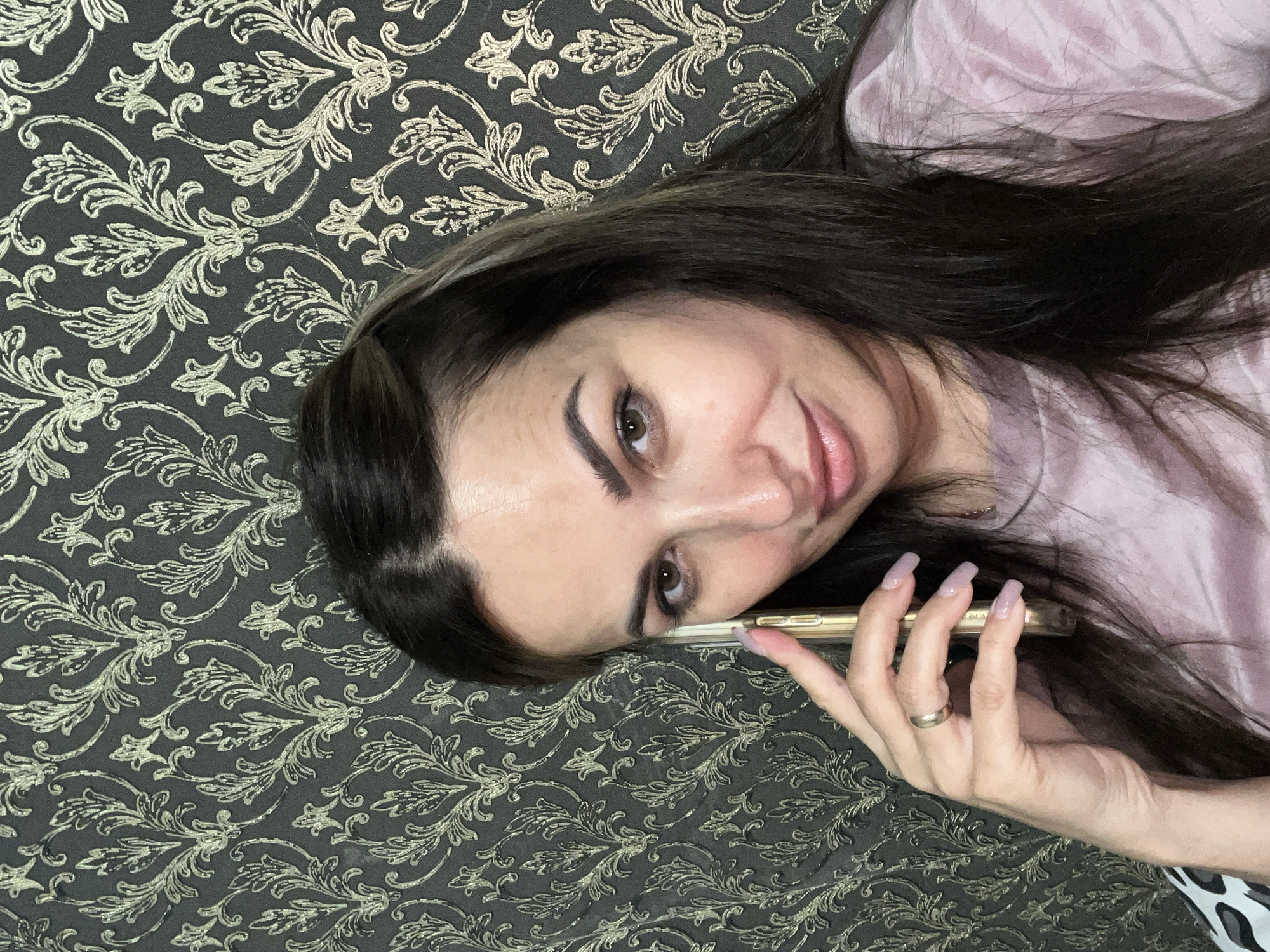 A medium shot selfie taken indoors shows a woman with long, dark hair, holding a gold-colored smartphone to her right ear. She is looking towards the camera with a slight smile. Her left hand is raised to her face, with her index finger near her cheek. She is wearing a light pink t-shirt. Behind her is a wall with dark gray wallpaper featuring a gold, ornate damask pattern. The lighting appears to be natural and soft.