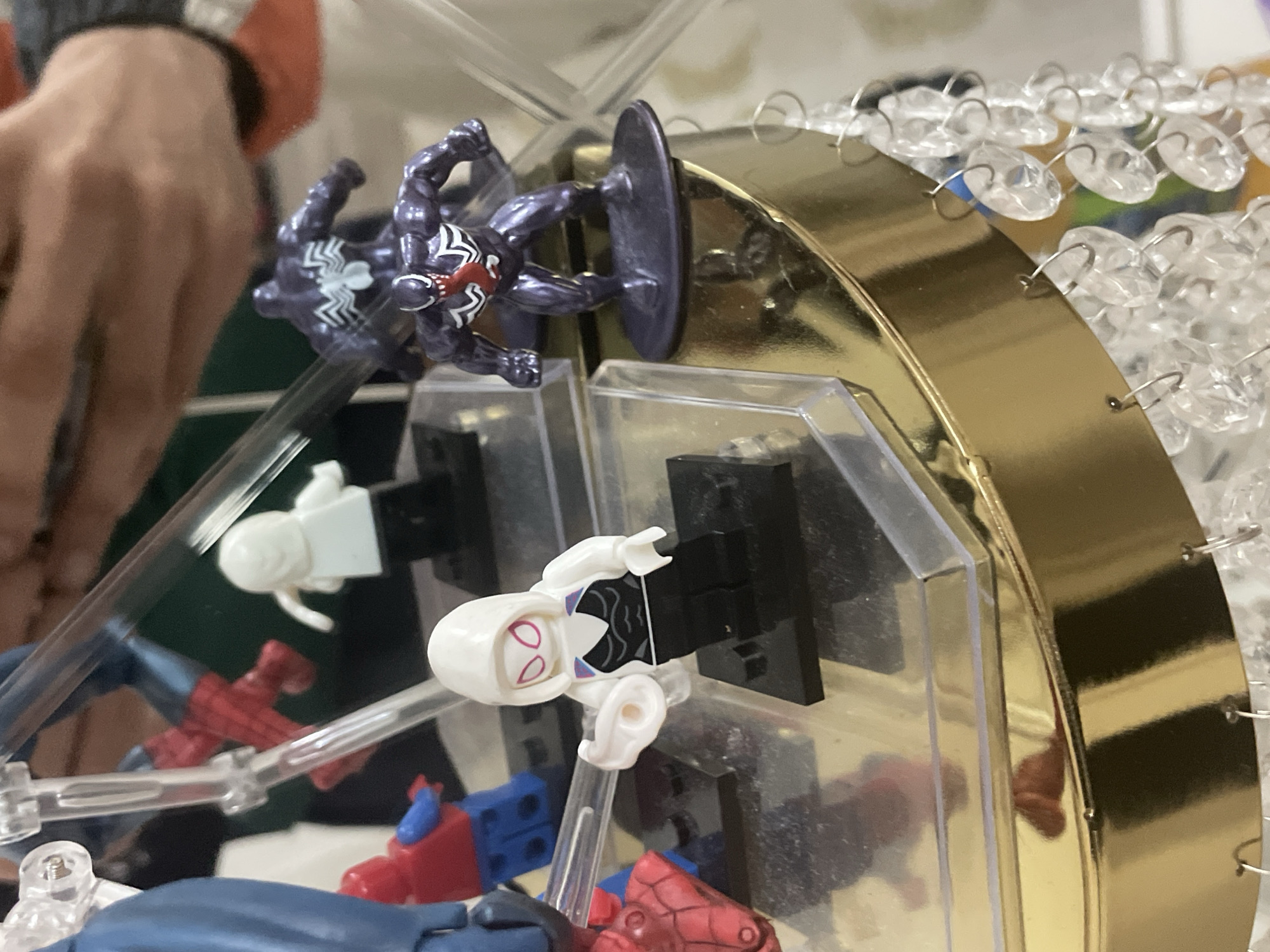 The image displays a close-up view of various Marvel-themed minifigures and action figures arranged on a multi-tiered, reflective display stand, located in the city of Ihtiman, Bulgaria. In the foreground, a Lego minifigure of Spider-Gwen, characterized by a white hood, black and white torso with pink accents, and black legs, stands on a black base, extending a white web-shooter accessory from her right hand. To her right, in the mid-ground, two dark purple Venom action figures are present; the front figure has a prominent white spider symbol on its chest and a red tongue protruding, standing on a dark circular base. The rear Venom figure, slightly out of focus, also displays a white spider symbol on its back. Behind Spider-Gwen, a partially obscured red and blue Lego minifigure (Spider-Man) is visible, along with a mostly white Lego minifigure on a black base. Further to the far left, part of a blue figure is seen. The figures are supported by clear plastic stands and are resting on a golden, highly reflective surface that forms the primary tier of the display. Below this gold surface, numerous clear, faceted decorative beads, strung on silver hooks, hang in a chandelier-like arrangement. The background is out of focus, revealing blurred elements including skin-toned hands in the upper right quadrant and indistinct colors of a domestic interior.