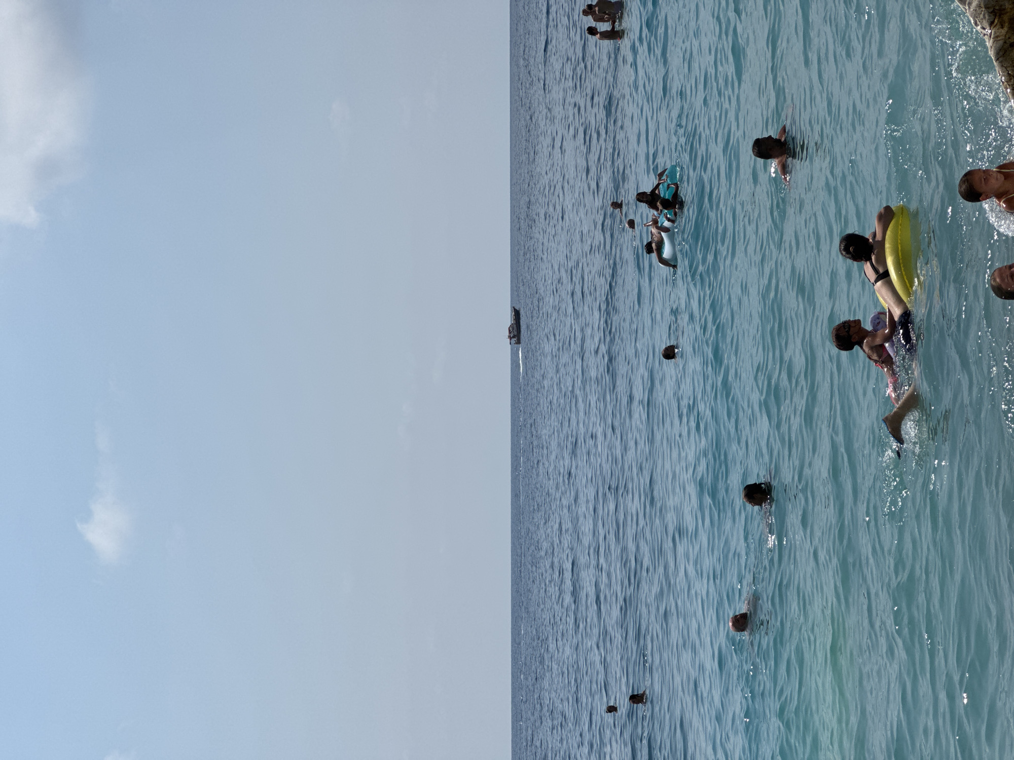 In the city of Mirište, Montenegro, a group of people are enjoying a sunny day at the beach. The crystal-clear turquoise water is calm, with gentle waves lapping against the shore. The sky is a pale blue with a few scattered white clouds, suggesting a pleasant weather day.
In the water, several individuals are visible. Some are swimming, while others are relaxing on inflatable tubes and rafts. A family can be seen enjoying a yellow inflatable ring, with two children and an adult in the water. In the distance, a small boat is navigating the sea. The overall scene conveys a sense of leisure and recreation, typical of a summer day at the beach.