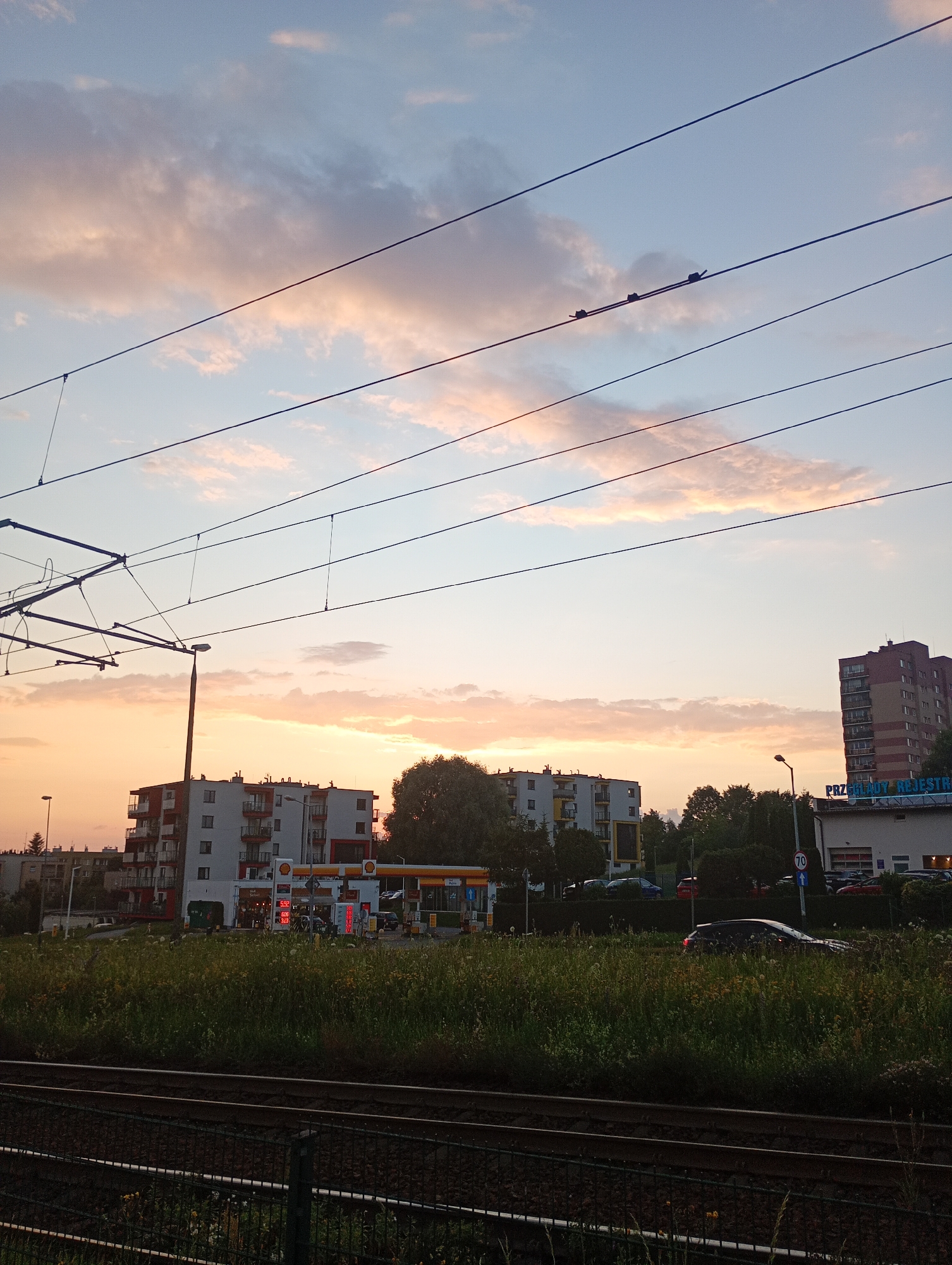 This is a medium shot, taken at eye level, of a city street scene at dusk in Krakow, Poland. The sky is a soft gradient of blue and pink, with scattered clouds that are illuminated by the setting sun. Multiple overhead power lines, likely for trams or trains, stretch across the frame, casting thin shadows.
In the middle ground, several multi-story apartment buildings are visible. One building, predominantly white with red accents and balconies, stands out, with a Shell gas station directly in front of it, complete with visible gas pumps and illuminated price displays. Another apartment building, similar in style but predominantly gray, is to the left. Beyond these buildings, trees and more distant structures can be seen.
To the right, a taller brick building with many windows rises above the others, with a sign that reads "PRZEGLĄDY REJESTRACJE". Further to the right, a smaller white building with a garage door is visible.
In the foreground, train tracks run horizontally across the bottom of the image, separated by a wire fence. A dark green, overgrown grassy area with wildflowers separates the tracks from the street and buildings. A black car is parked in this grassy area.
The overall mood is peaceful and quiet, typical of a city at the end of the day. The soft lighting suggests it is either sunrise or sunset, and given the colors, sunset is more likely. The presence of apartment buildings, a gas station, and the sign for vehicle inspections point to an urban or suburban environment.