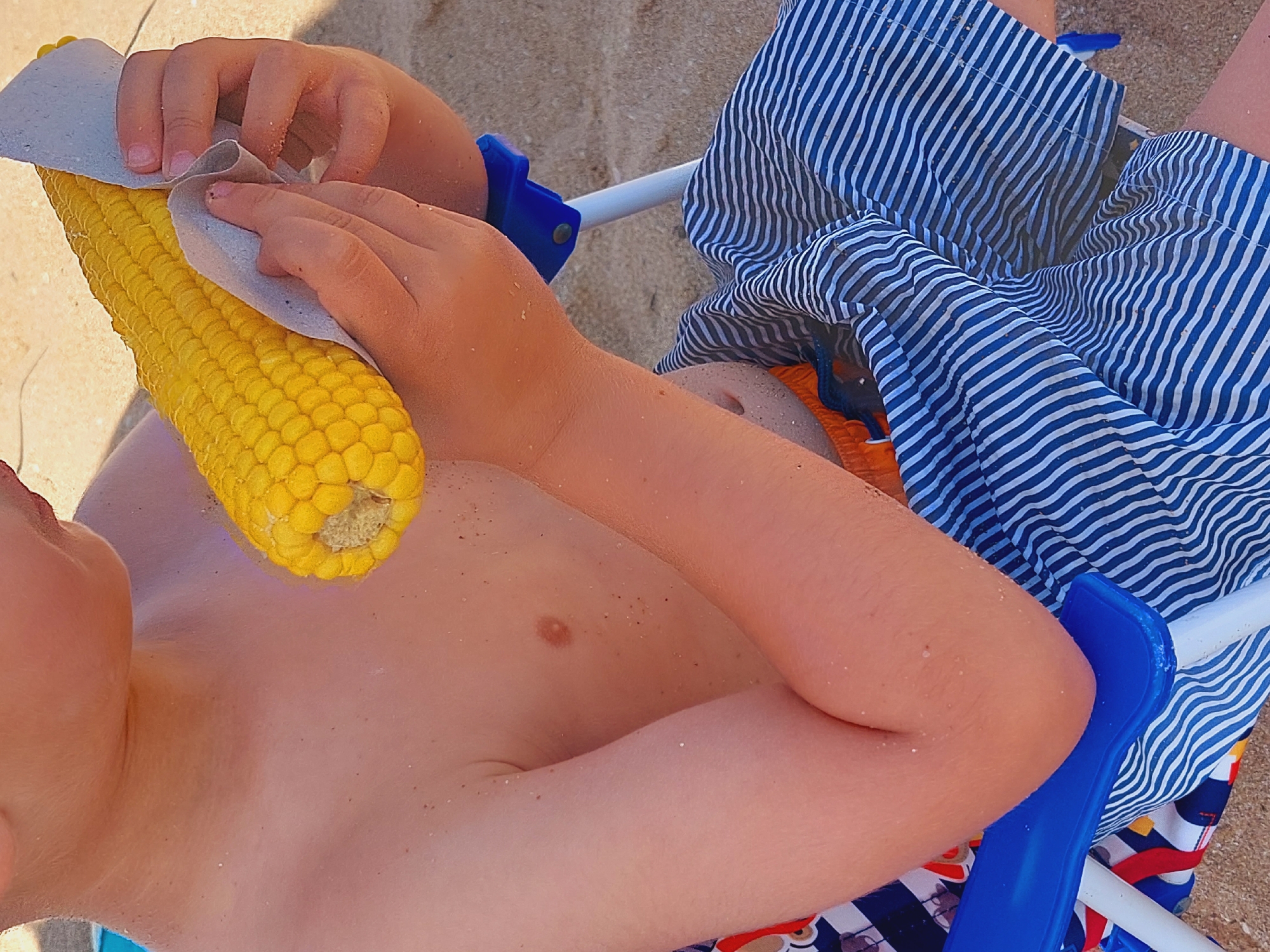 This media file shows a close-up, overhead view of a young person on a sandy beach. The person is lying down, likely on a beach chair, as evidenced by a blue plastic chair support and a striped towel or beach blanket in blue and white. The person is shirtless and appears to be in swimwear, with part of orange swim trunks visible.
The main activity captured is the person holding a cob of corn with both hands. The corn is bright yellow and appears to be raw, with the kernels clearly visible. The person's hands are a light skin tone and are gently holding the corn, with one hand partially covered by a light gray cloth or towel. A small amount of sand is visible on the person's skin and on the corn.
The setting is a beach, indicated by the sand. The lighting suggests it is daytime, possibly sunny, given the bright colors and the contrast. No specific time of day or weather conditions are explicitly evident beyond it being a bright day. There is no visible text in the media file. The overall mood appears relaxed and casual, typical of a day at the beach. The location context of Sozopol, Bulgaria, places this scene in a coastal town.