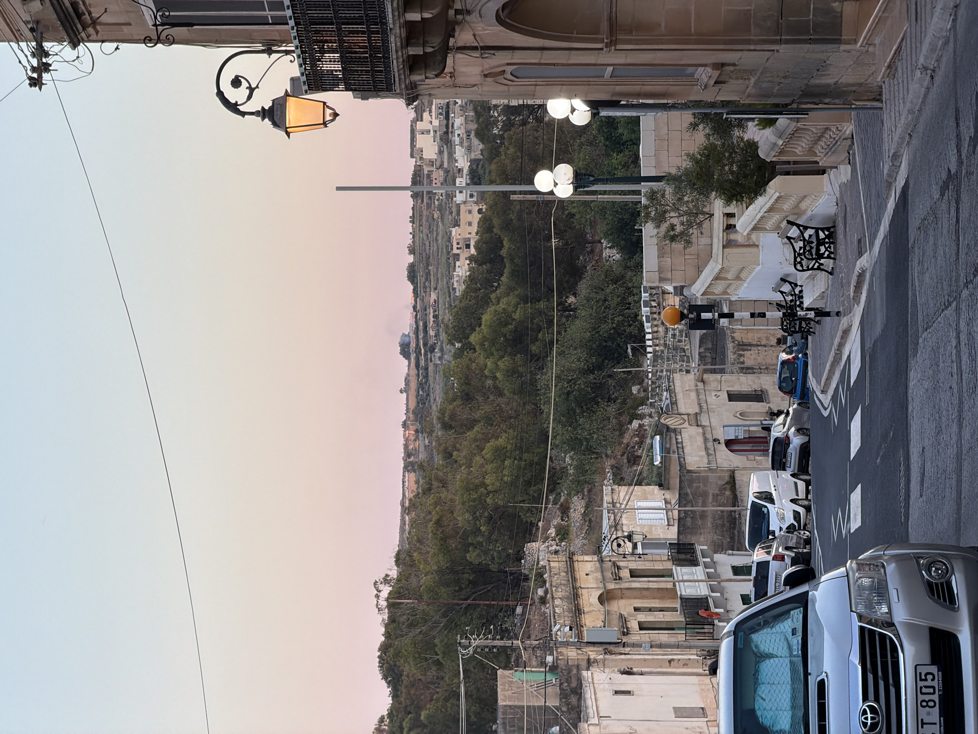 The image depicts a street scene in Fontana, Malta, at dusk. A street lamp with an illuminated amber bulb is visible on the right side of the frame, attached to a building with a balcony. In the foreground, the front of a silver SUV is visible, parked facing away from the camera. The street slopes downwards, with buildings lining both sides. Several other vehicles are parked or driving on the street, including a white van, a silver SUV, and several cars. A pedestrian crossing is marked on the road. In the background, trees and a distant landscape with buildings are visible under a sky transitioning from pale blue to soft pink. The architecture suggests a Mediterranean style with stone buildings and balconies.