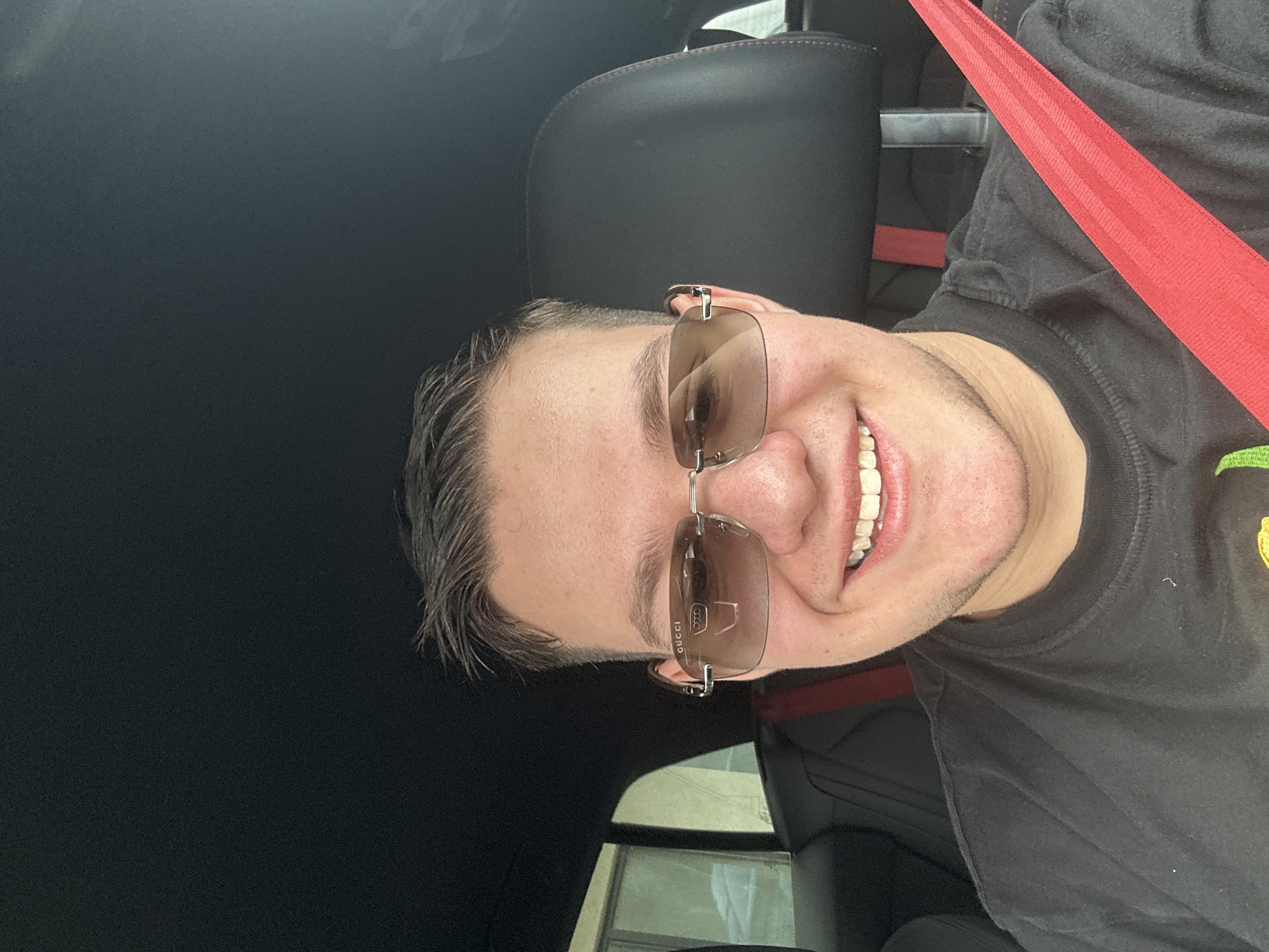 A man is sitting in a car, wearing sunglasses and a black t-shirt with a red seatbelt. The car has black leather seats with red stitching. The interior of the car is black. The outside environment is visible through the window. The location in the city of Sofia, Bulgaria cannot be determined.