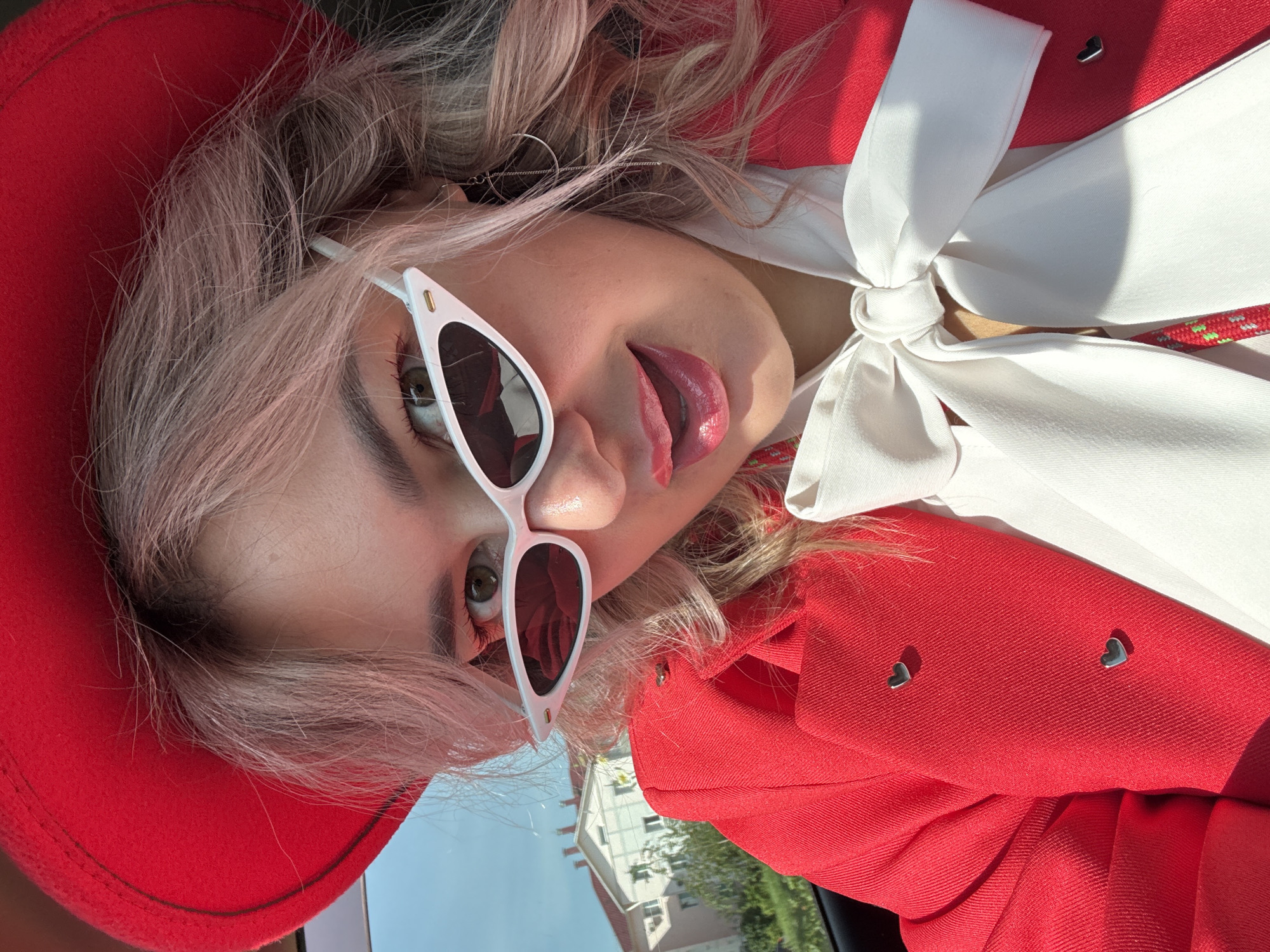 A close-up, selfie-style photograph features a young woman with light pink hair, wearing a red hat, white cat-eye sunglasses, and a bright red blazer adorned with two small heart-shaped pins. She has on a white bow-tie blouse underneath the blazer, and her lips are painted with glossy pink lipstick. The background suggests an outdoor setting with a clear blue sky, a building with a red roof, and green foliage. The overall impression is one of a stylish and vibrant individual, possibly in Birobidzhan, Russia, captured in a moment of playful self-expression.