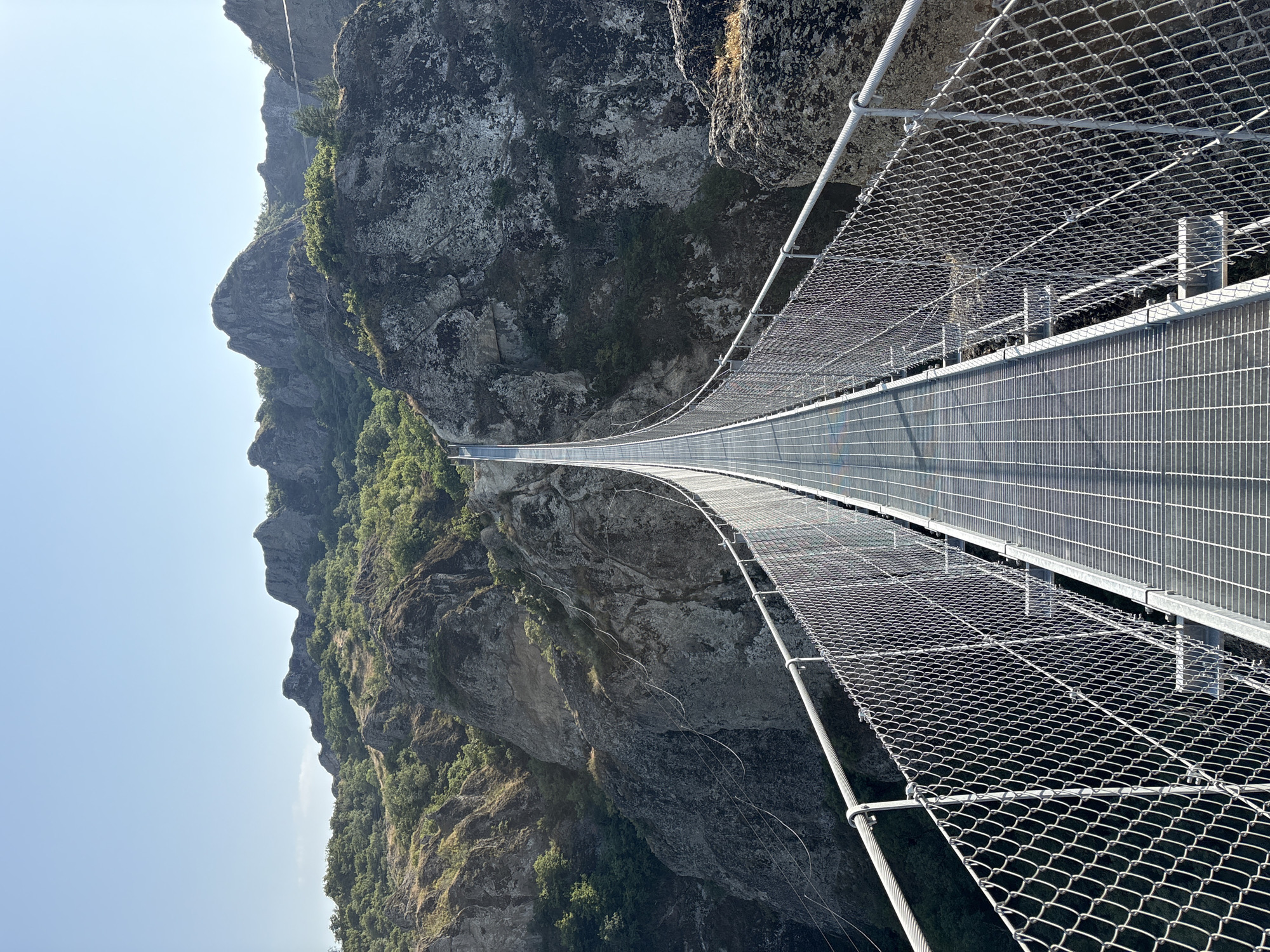 The image depicts a long, narrow suspension bridge stretching across a rocky, mountainous canyon in Orbeti, Georgia. The bridge is constructed with a metal grate walkway and is enclosed by chain-link fencing and thick cables. The surrounding landscape is rugged and arid, with steep rock faces and sparse vegetation. The sky above is clear and blue. There are no people visible in the image.