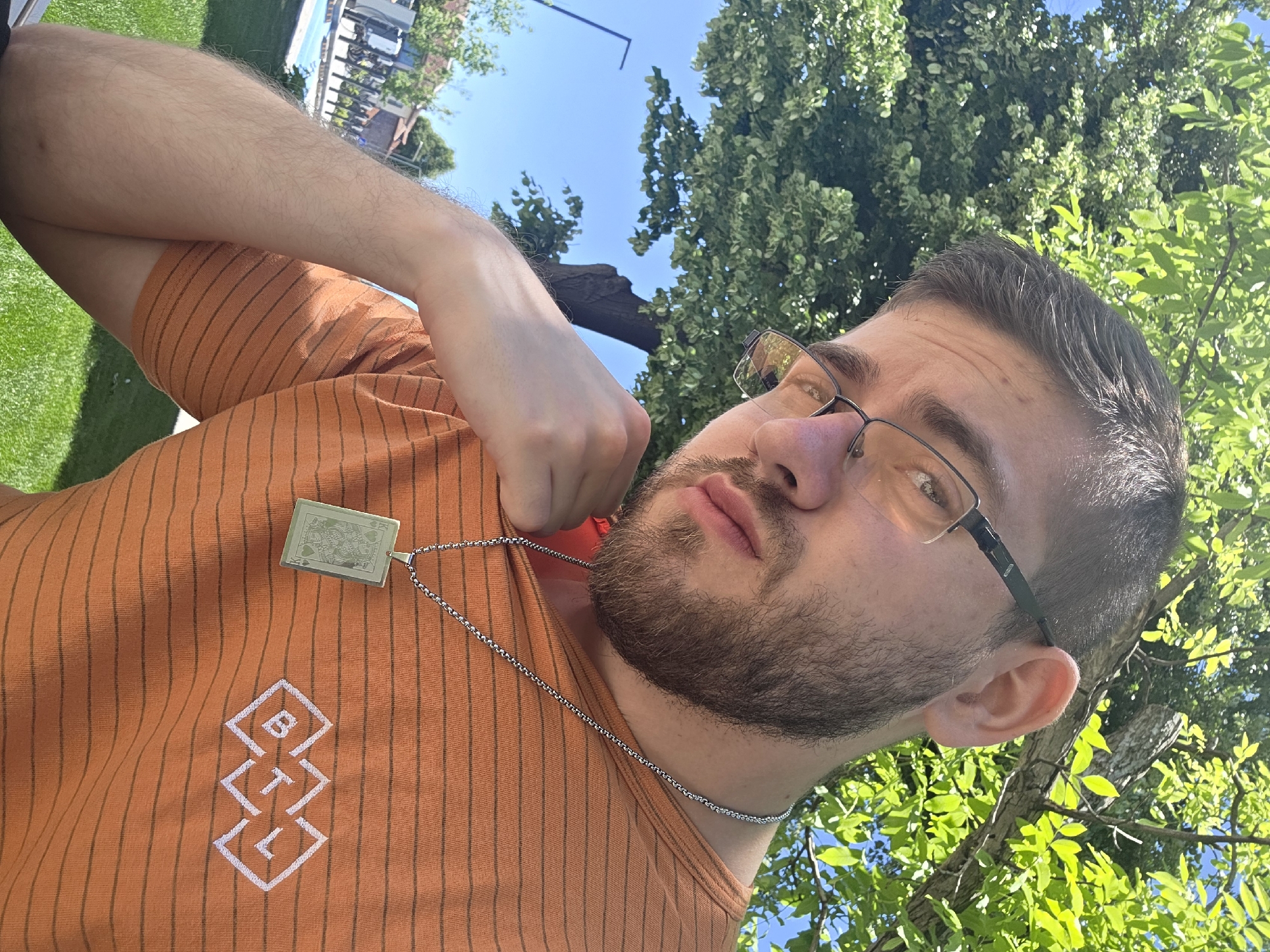 A man is lying down, facing upwards towards the sky, with trees and blue sky visible in the background. He wears glasses and an orange striped shirt with the white letters "BTL" arranged vertically in diamond-shaped outlines on the left side of the chest. A silver chain necklace with a rectangular pendant, displaying what appears to be a card, is visible around his neck. The man is outdoors, likely in a park or yard with grass and trees. The location cannot be specifically determined within the city of Plovdiv, Bulgaria, without additional information.