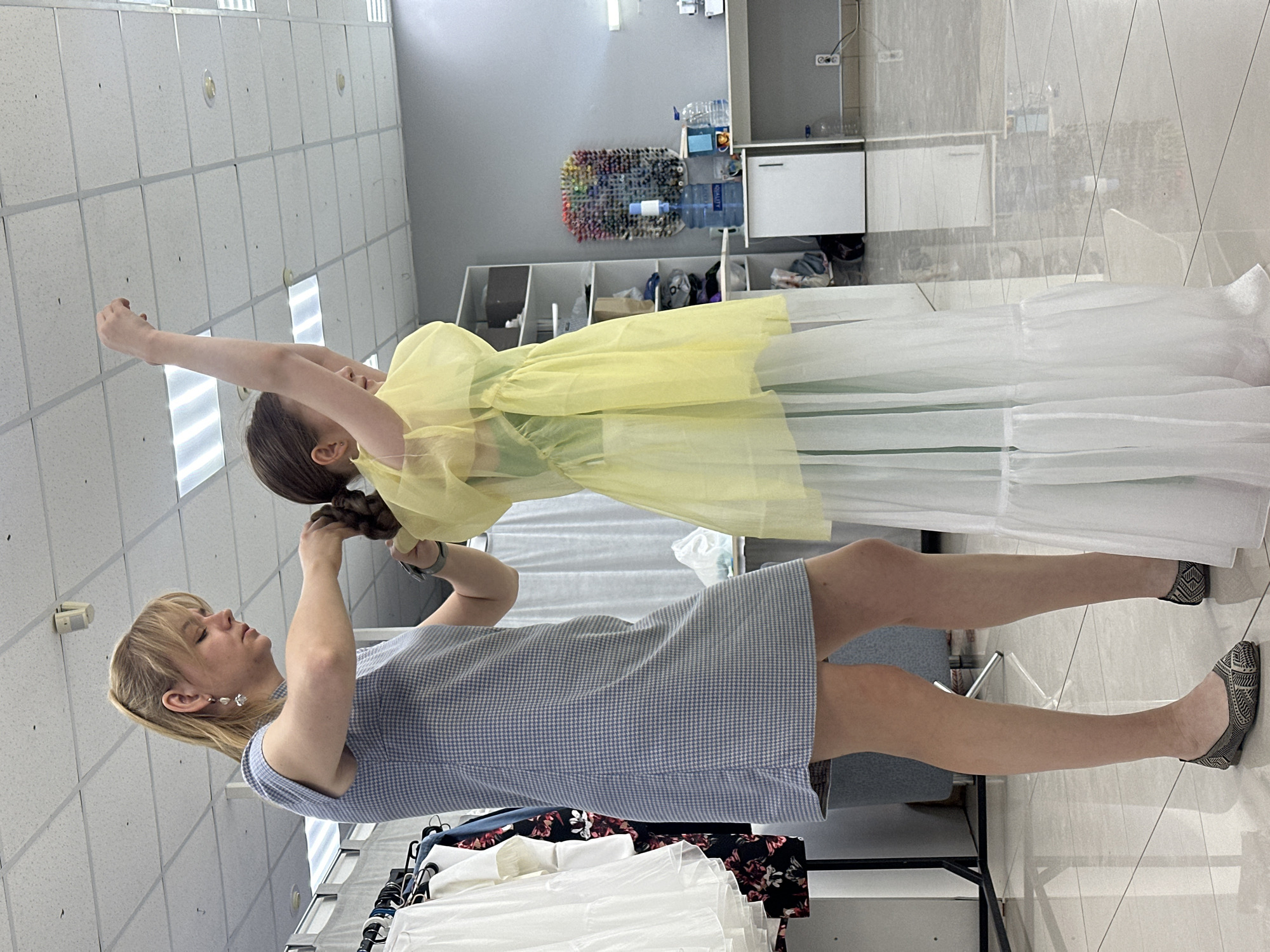 Two women are interacting in a brightly lit indoor space. The first woman, on the left, wears a blue and white checkered dress and silver flats. She is holding the hair of the second woman, who is wearing a yellow and white dress. The scene includes a clothes rack, shelving units, and other furniture. A grid-patterned ceiling with recessed lighting is visible above. The location is likely a dressmaking or design studio in Votkinsk, Russia.