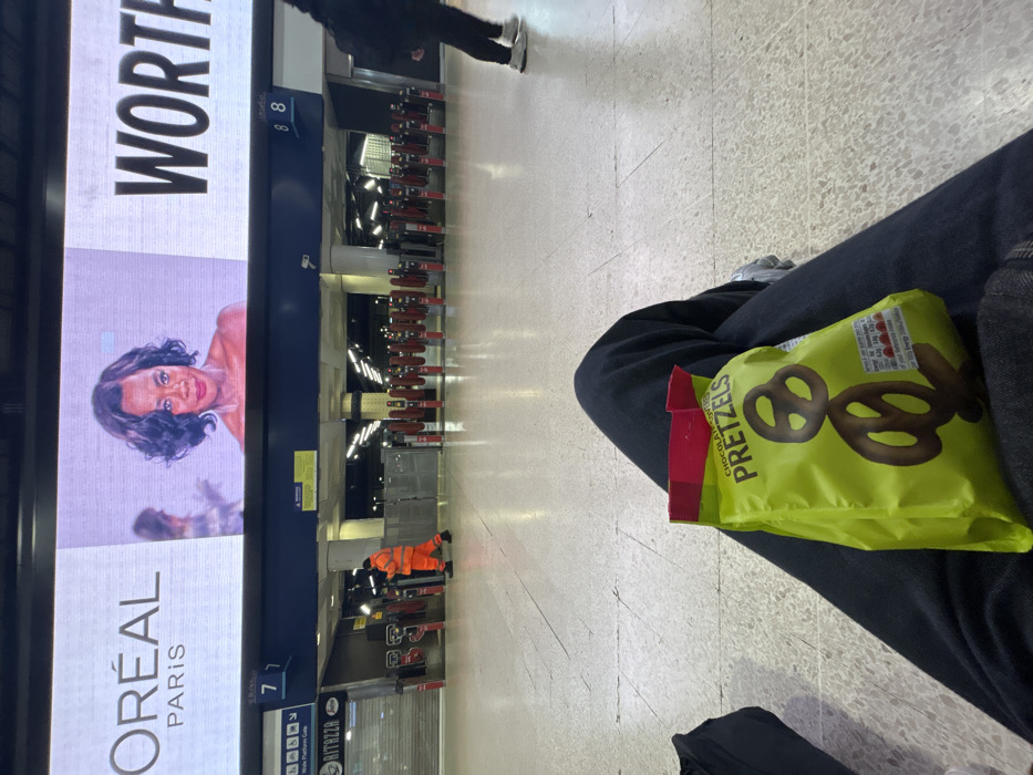 The image depicts an interior view of London Waterloo railway station, United Kingdom. In the immediate foreground, a person's upper thighs are visible, clad in dark trousers, with a bright green, open bag of "Chocolate Covered PRETZELS" resting on their lap. A black bag is partially visible to the left of the legs. The floor is composed of large, light-colored square tiles.
In the midground, a row of red and grey ticket barriers (turnstiles) extends horizontally across the width of the frame. To the left of these barriers, a retail unit with a closed metal roller shutter is visible, bearing the name "RITAZZA." A person in an orange high-visibility uniform and dark shoes is seen walking from right to left, passing near the left side of the ticket barriers. Another person, dressed in dark clothing (black jacket, dark trousers, white trainers), is partially visible on the far right, walking leftwards.
Above the ticket barriers, a large digital display screen dominates the background. The screen displays an advertisement: on the left, "L'ORÉAL PARIS" is visible, and prominently in the center is a headshot of a woman with dark, wavy hair, looking forward, likely Viola Davis. To the right of her image, the partial word "WORTH" is displayed. Below this main screen, a blue fascia panel indicates platform numbers "7" (with "Platform Gate" text) and "8." A surveillance camera is mounted on a pillar beneath this panel. The overall lighting suggests an indoor, brightly lit public space.