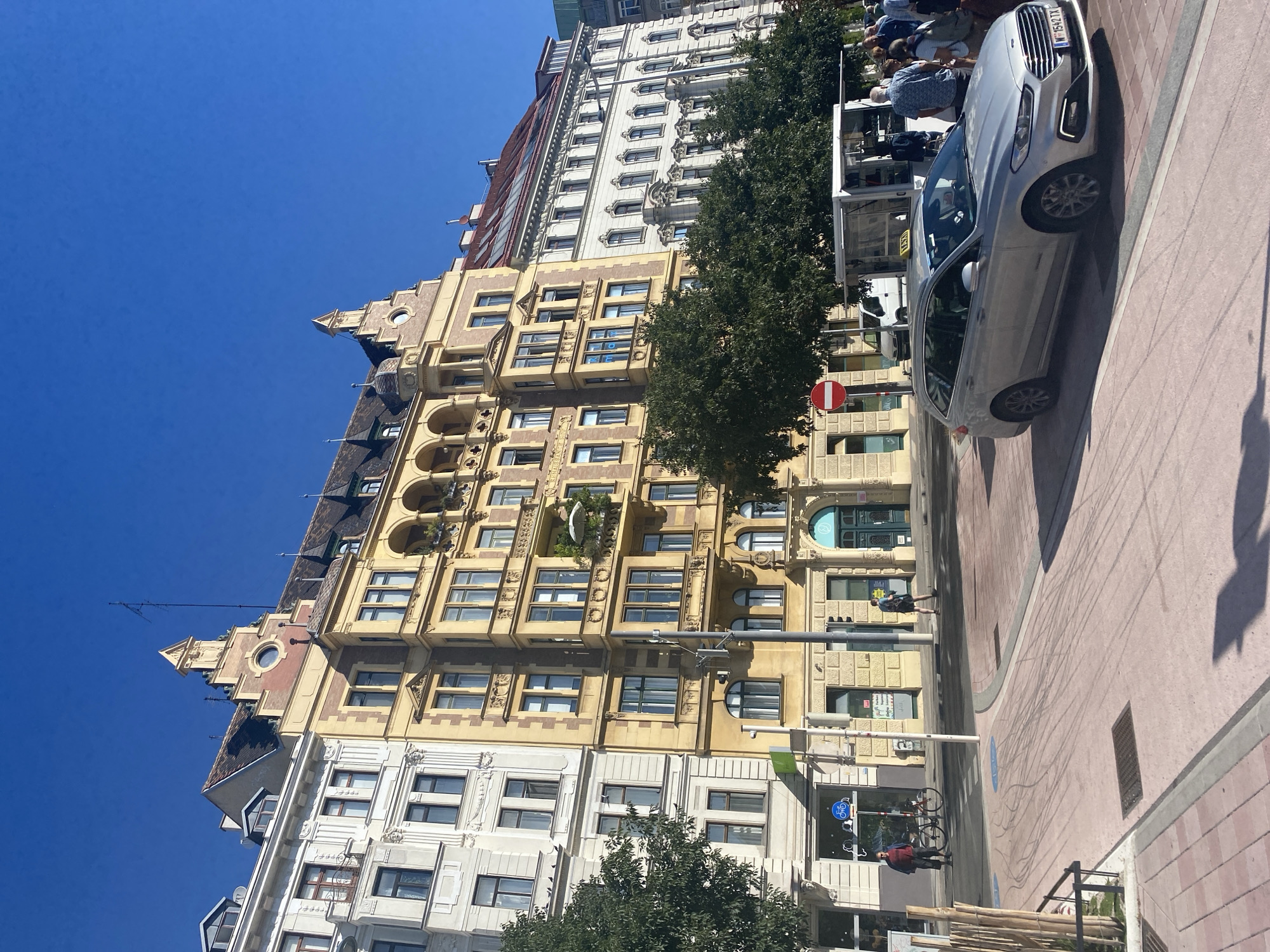 The image depicts a sunny street scene in Vienna, Austria, with a row of ornate, multi-story buildings lining the right side. The architecture is diverse, featuring facades of white, yellow, and a red-tiled roof. The buildings showcase classical elements such as balconies, arched windows, and decorative cornices. In the foreground, a silver station wagon with a taxi sign on its roof is parked. People are visible in the background, some walking, others standing near a white kiosk-like structure. Trees with dense green foliage are interspersed along the street. The ground is paved with pinkish-grey tiles, and there are tire marks on the pavement, suggesting recent vehicle activity. A red and white circular traffic sign with a horizontal bar is mounted on a pole to the left. The sky is clear and bright blue.