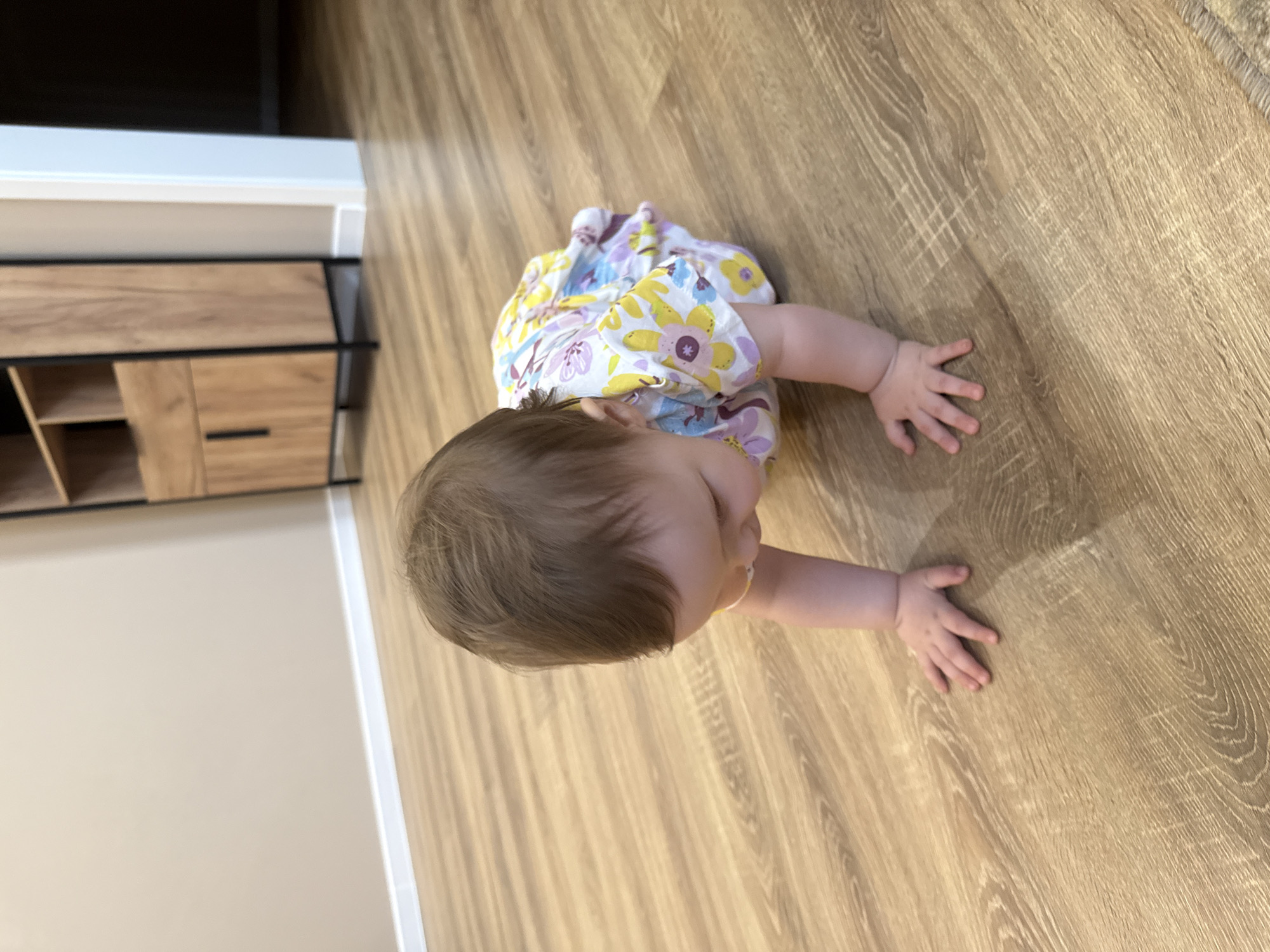 A baby is crawling on a wooden floor. The baby is wearing a dress with a floral pattern. The scene appears to be indoors, possibly in a home. The lighting suggests it could be daytime. There are no discernible emotions or specific activities beyond crawling. No text is visible in the image.