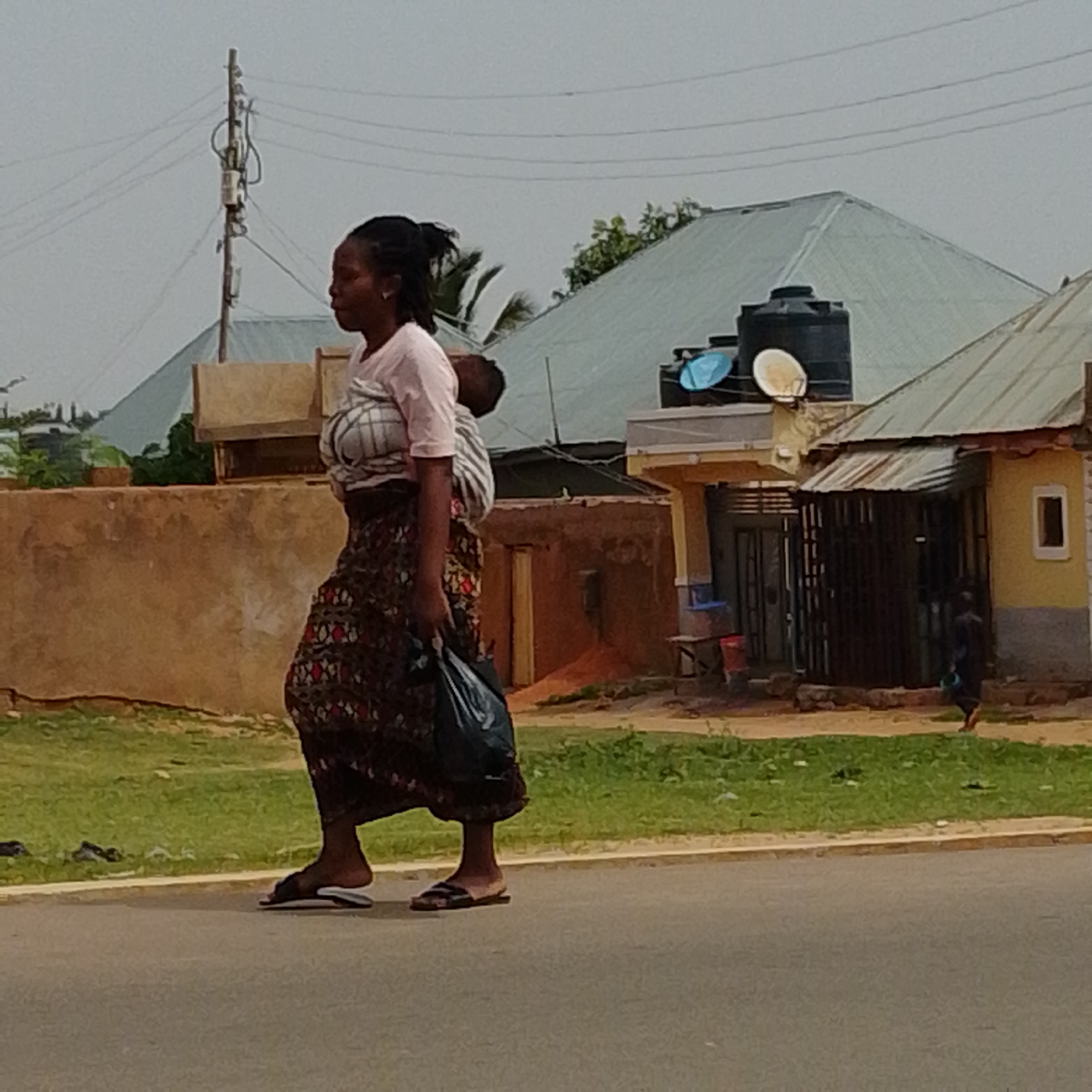 A woman walks on a paved road in an outdoor, urban setting. She is wearing a pink shirt and a dark, patterned skirt, and she is carrying a baby wrapped in a cloth on her back. She also holds a black plastic bag in her left hand. The background shows buildings with metal roofs, some with water tanks and satellite dishes, and a telephone pole with wires. The ground is covered with green grass and dirt.
