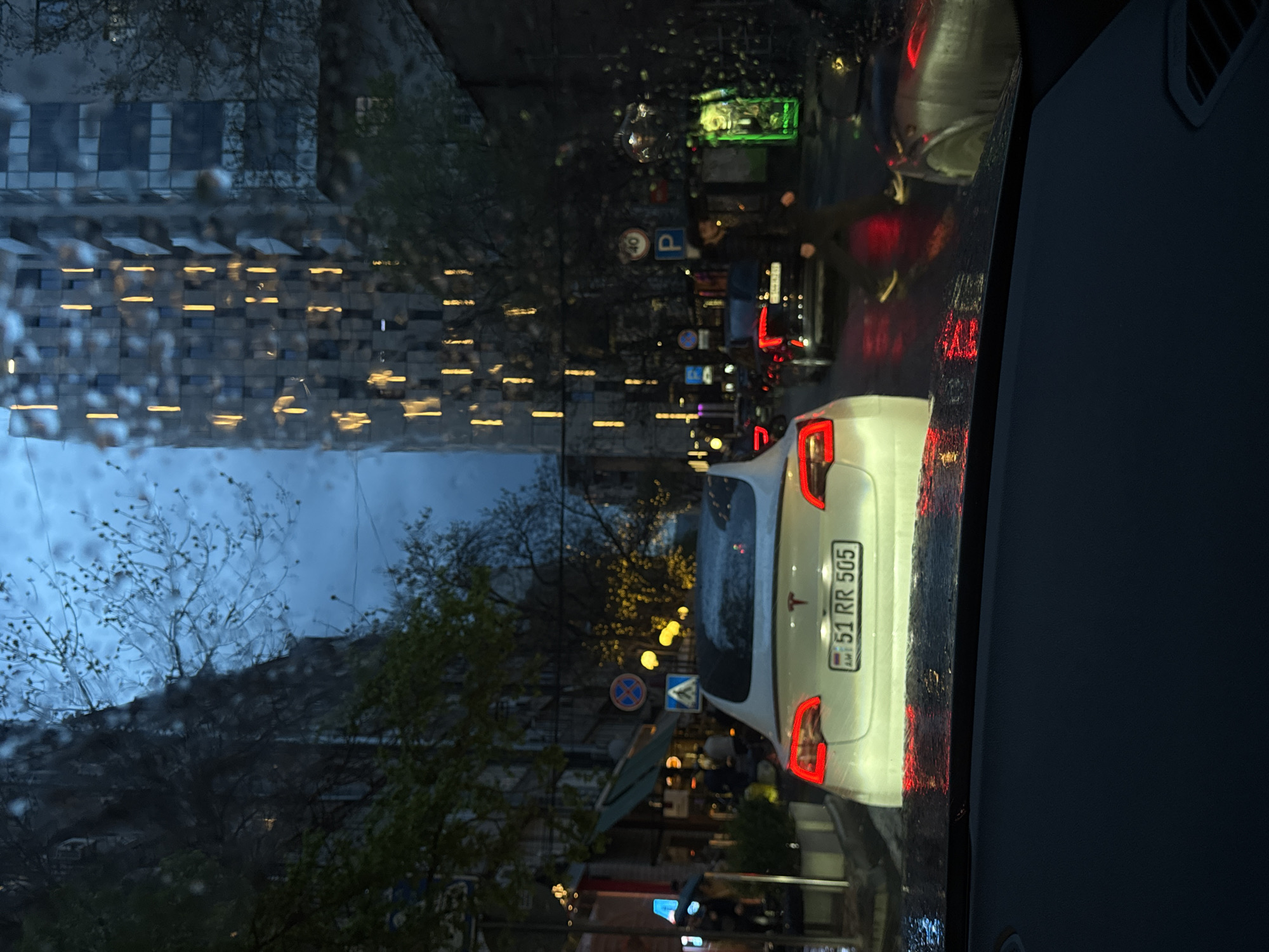 The image shows a view from inside a car, likely the driver's perspective, on a rainy evening in Yerevan, Armenia. The car is positioned on a city street with a white Tesla in front, displaying the license plate "51 RR 505". The city skyline is visible through the rain-streaked windshield, including tall buildings with illuminated windows. Street lights and traffic signals are present, as well as pedestrian and traffic signs. Another vehicle with illuminated taillights is also in view behind the Tesla. Pedestrians are present on the sidewalk. The overall environment suggests a typical urban setting during inclement weather.