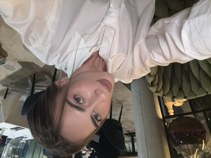 A woman poses for a selfie while sitting at a table. She has light brown hair and blue eyes, and she is wearing a white button-down shirt. An olive-green quilted couch is behind her. In the background, there is an elaborate decorative glass sphere sitting on a stand and a patterned tile floor. The photo is taken from a slightly high angle.