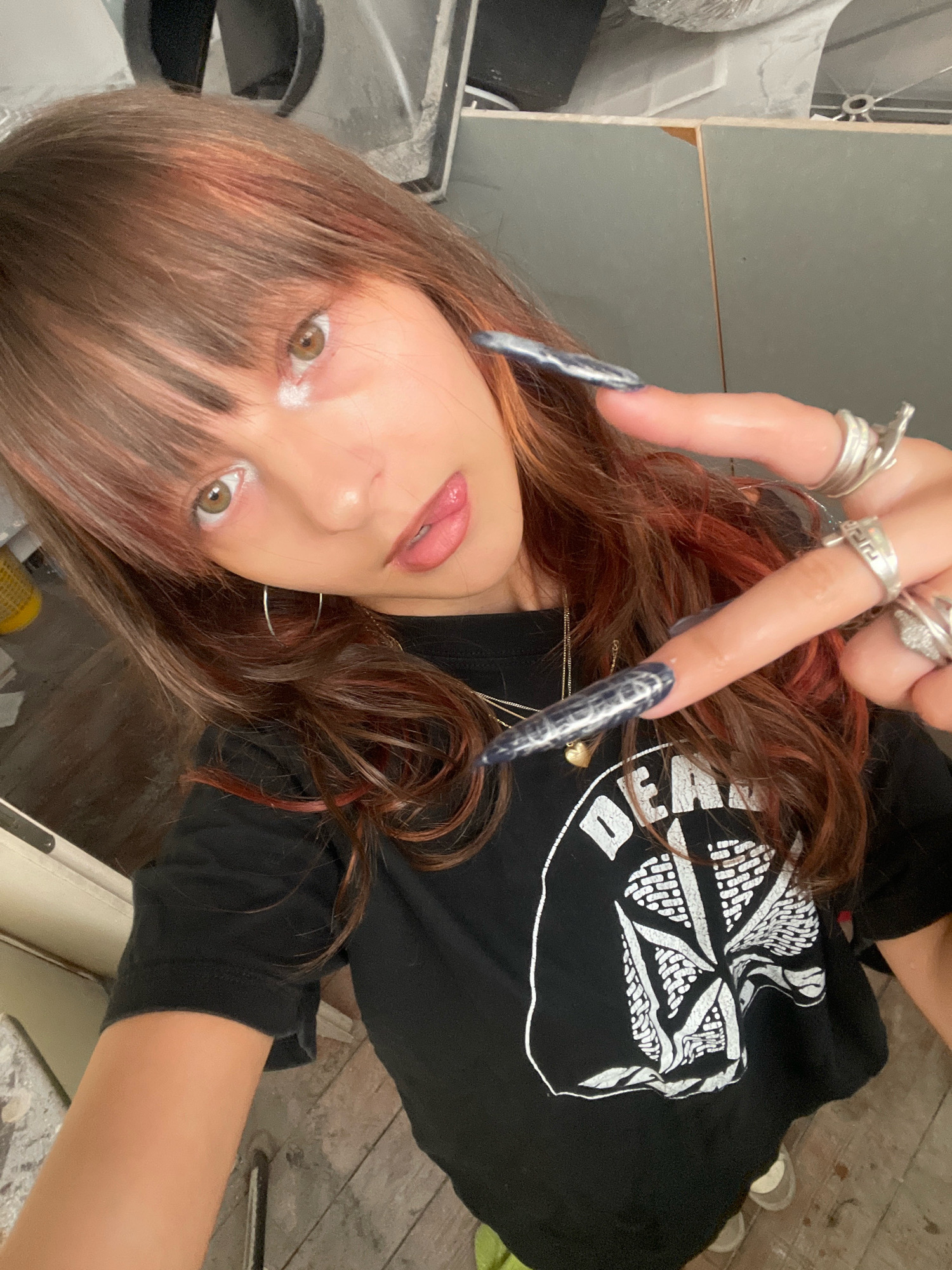 A young woman with long, brown hair, featuring streaks of red and blonde, is pictured in a selfie. She is wearing a black t-shirt with a white graphic design. Her eyes are wide open, and she has light makeup on, including white eyeshadow and pink lipstick. She is adorned with several silver rings on her fingers and has long, dark, pointed fingernails. She is holding up two fingers in a peace sign towards the camera.
The background suggests an indoor setting with wooden flooring and what appears to be a wall constructed of gray panels or possibly unfinished building materials. There are also glimpses of what might be a window or a doorway. The overall environment seems somewhat utilitarian, possibly a workshop or an unfinished room. The image does not contain any recognizable landmarks or specific details that would place it in the city of Raanana, Israel.