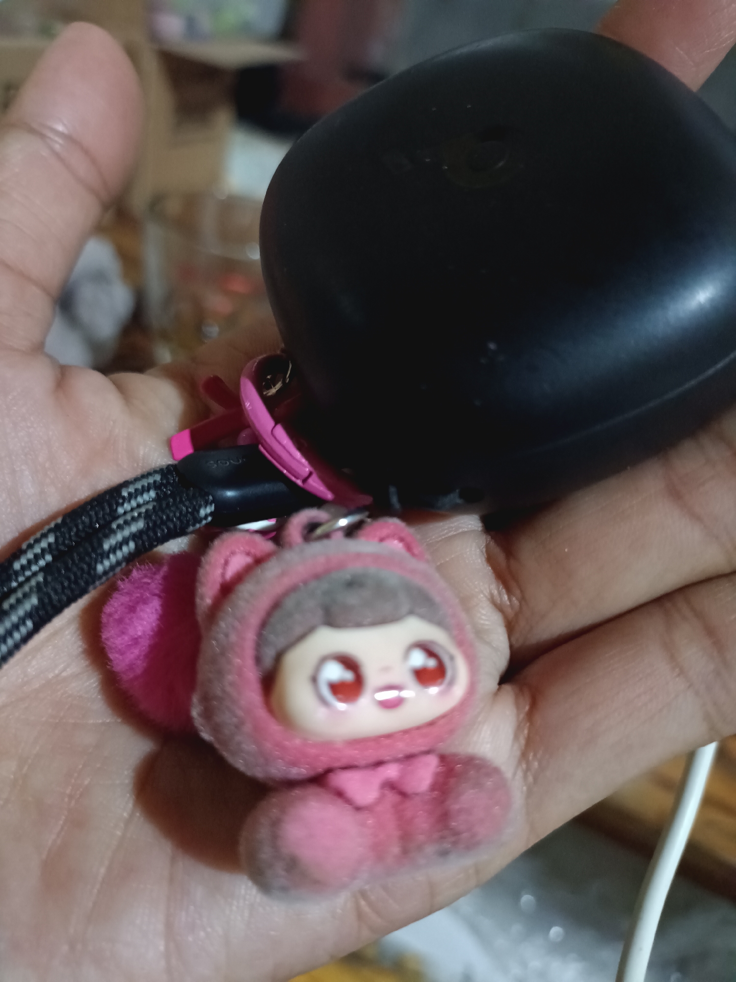 A close-up shot shows a hand holding a black charging case for earbuds and a fuzzy pink keychain of a doll with big red eyes. The doll is wearing a pink hooded outfit with a bow tie. A black and white braided lanyard is attached to the charging case and the keychain. The background is blurred, but appears to be an indoor setting with some boxes and a glass. The location is Ploso, Indonesia.