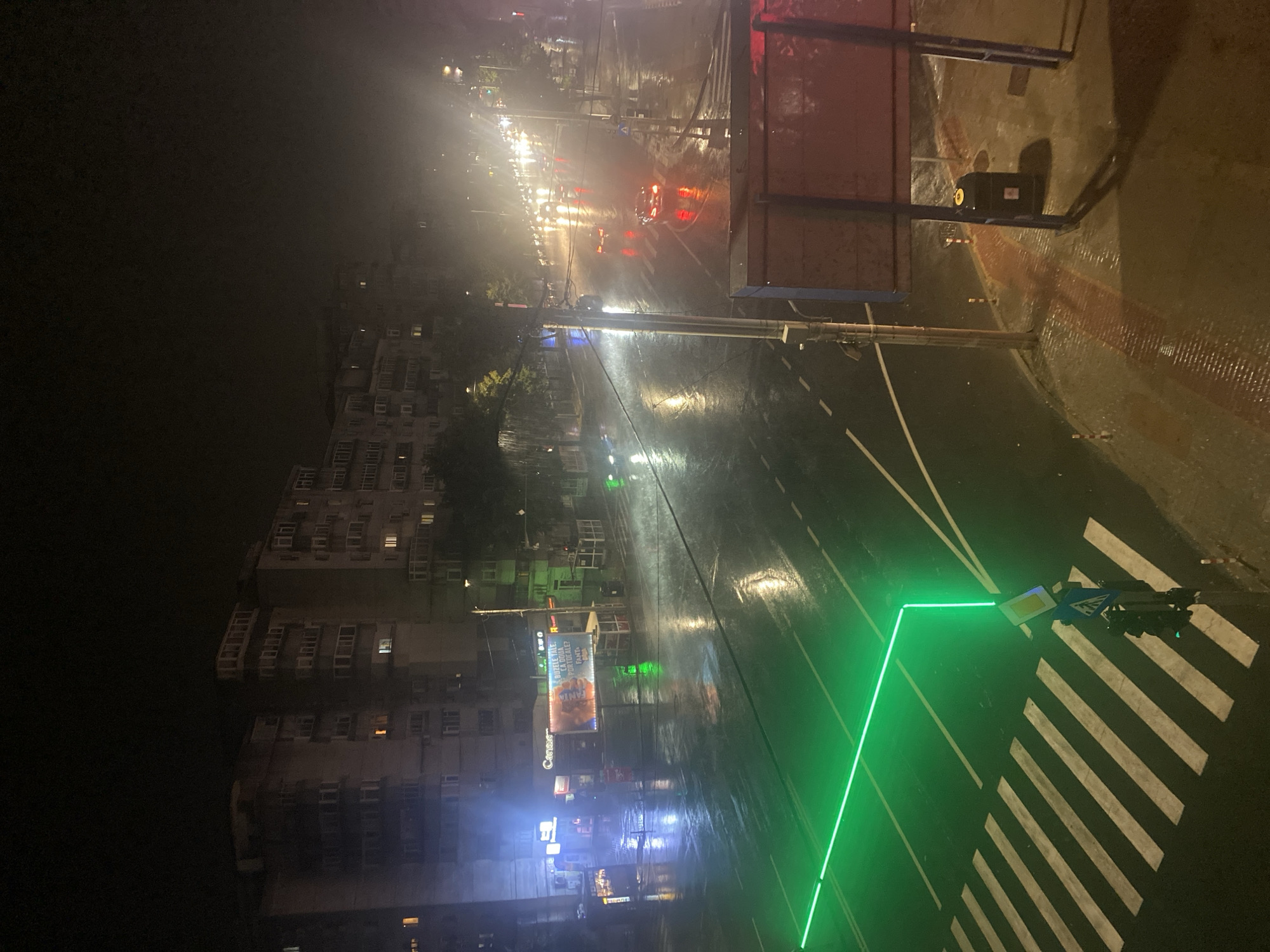 The image depicts a rainy night scene in Iași, Romania, likely on a street with apartment buildings on the left. A pedestrian crossing is prominent in the foreground, illuminated by a bright green line of light that outlines its edges and extends across the wet asphalt. Multiple cars are visible on the street, their headlights and taillights reflecting on the wet surface, creating streaks of light. The street appears to be a multi-lane road with traffic moving in both directions. In the background, several tall apartment buildings with illuminated windows are visible. There are also streetlights and shop signs contributing to the ambient light. A trash can and a bus stop shelter are situated on the right side of the image. The overall atmosphere is dark and wet, characteristic of a rainy evening.