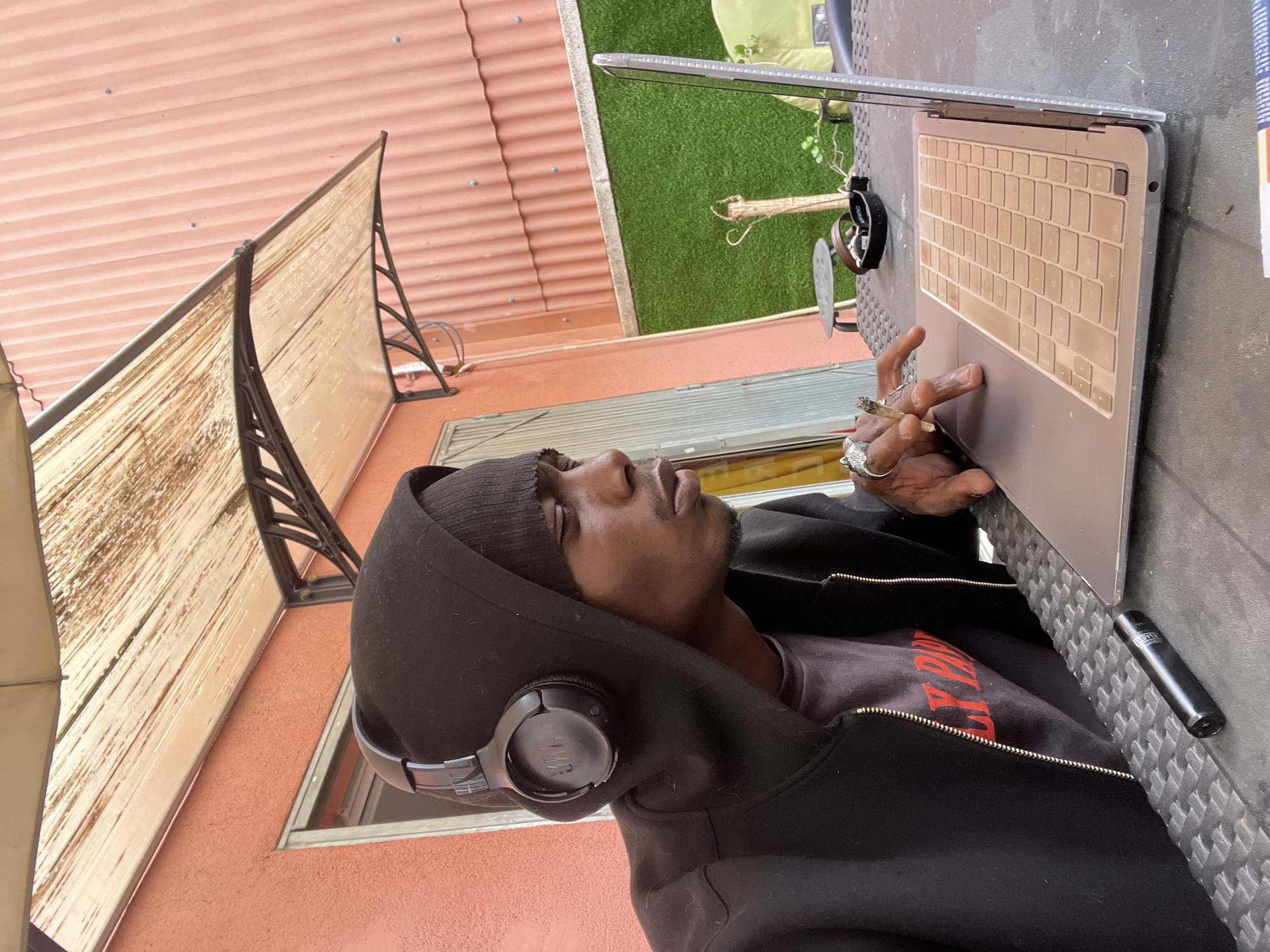 A man is depicted sitting outdoors, focused on a laptop. He wears a black hoodie with the hood up, a black beanie, and black JBL headphones. A dark t-shirt with partial red text is visible underneath his unzipped hoodie. He has several rings on his fingers, one appearing to be a silver animal head. In his right hand, he holds a lit cigarette or joint, with his thumb resting near the laptop's trackpad.
The laptop, silver in color and resembling an Apple MacBook, is placed on a dark, textured surface, possibly a patio table. Next to the laptop, a black Clipper brand lighter is visible.
The background suggests an outdoor setting, likely a balcony or patio. To the left, a translucent light-colored screen or divider with a dark frame is visible, partially obscuring a corrugated reddish-orange wall or roof structure. Behind the laptop and table, there's artificial green grass, a small plant, and what appears to be a light fixture or metal railing. The lighting indicates it's daytime, with a generally bright, diffused quality. The man's expression is neutral and concentrated on his activity.