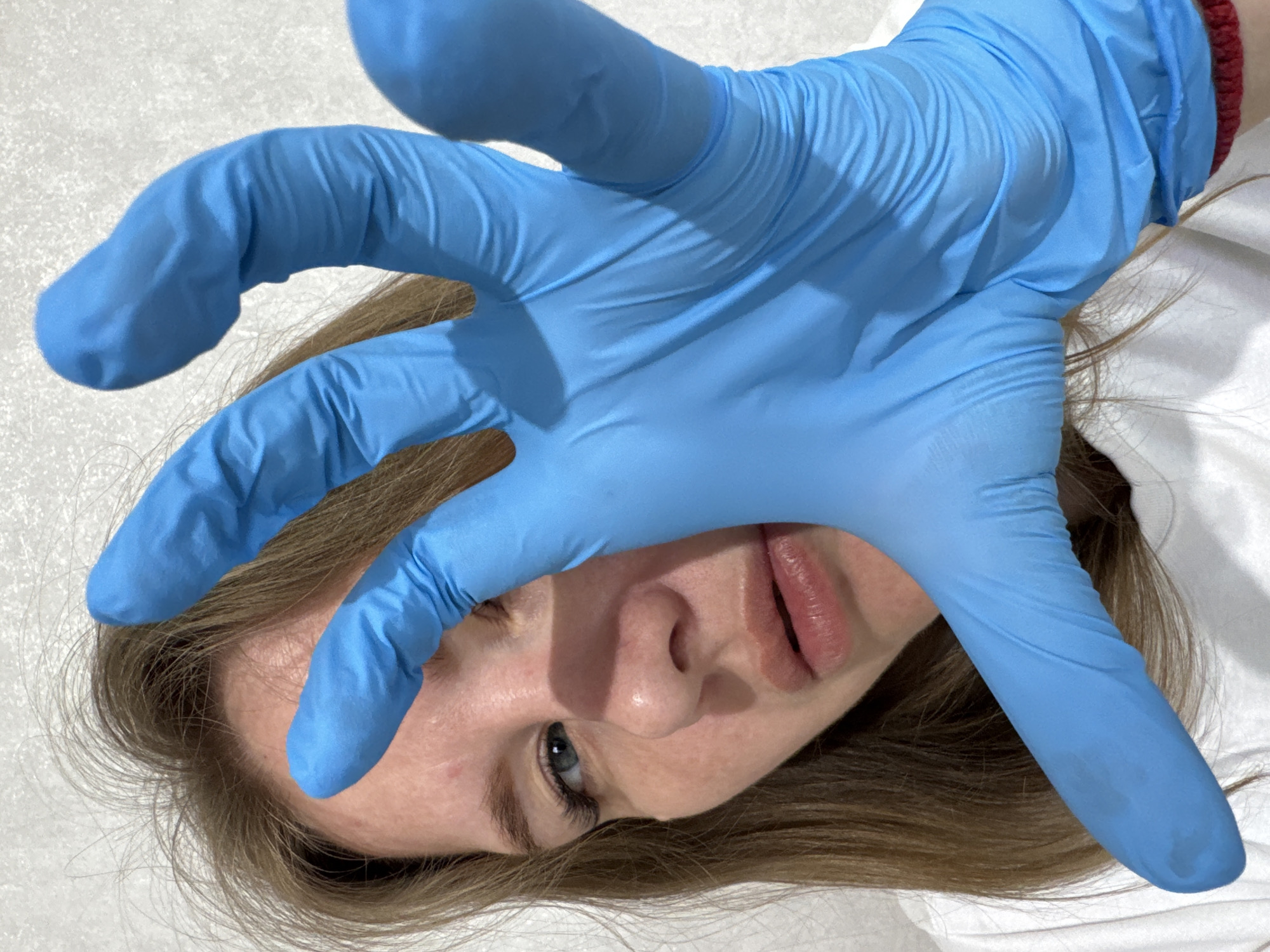A person lies on a white surface, likely a table or bed, in an indoor setting. The person has light brown hair and fair skin. A blue latex glove covers much of the person's face, obscuring the forehead and cheek. The person's mouth and nose are visible, along with one blue-grey eye. The red trim of a garment is visible under the glove, near the wrist. No specific details indicate a location in Gornyi, Russia.