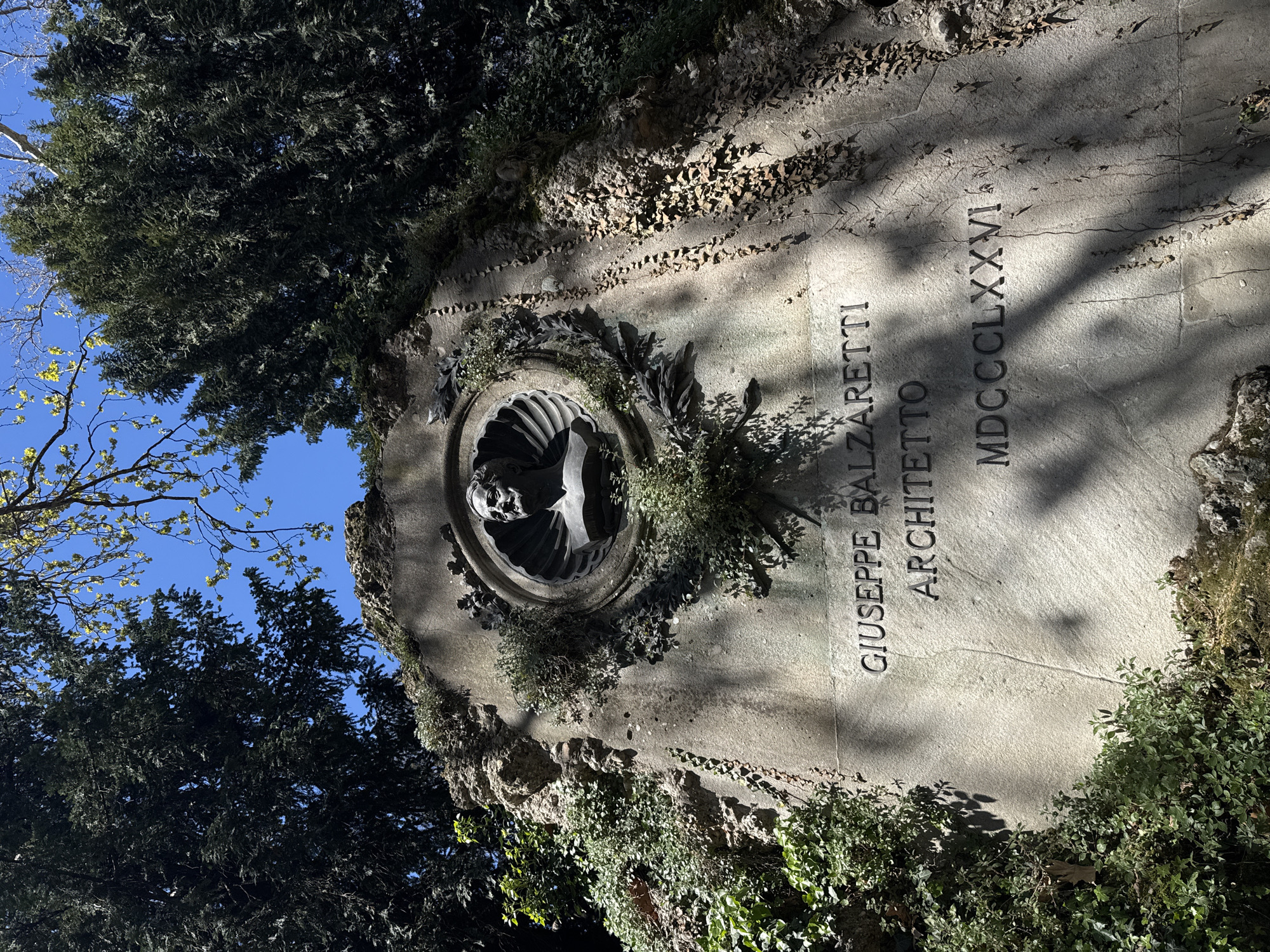 A medium-shot, low-angle photograph depicts a marble grave site in a Milan, Italy cemetery. The rectangular stone has a circular portrait, and vegetation is growing on the stone and surrounding area.
At the grave site's center is a dark, circular emblem with a bust in it. Foliage surrounds the emblem. To the emblem's right, text carved into the stone reads: "Giuseppe Balzaretti Architetto MDCCCLXXVI." Vegetation is growing on the grave site, especially near its edges, and dead leaves are scattered around it.
The grave site is surrounded by trees. The sky is blue.
The grave is for Giuseppe Balzaretti, architect, who died in 1876.