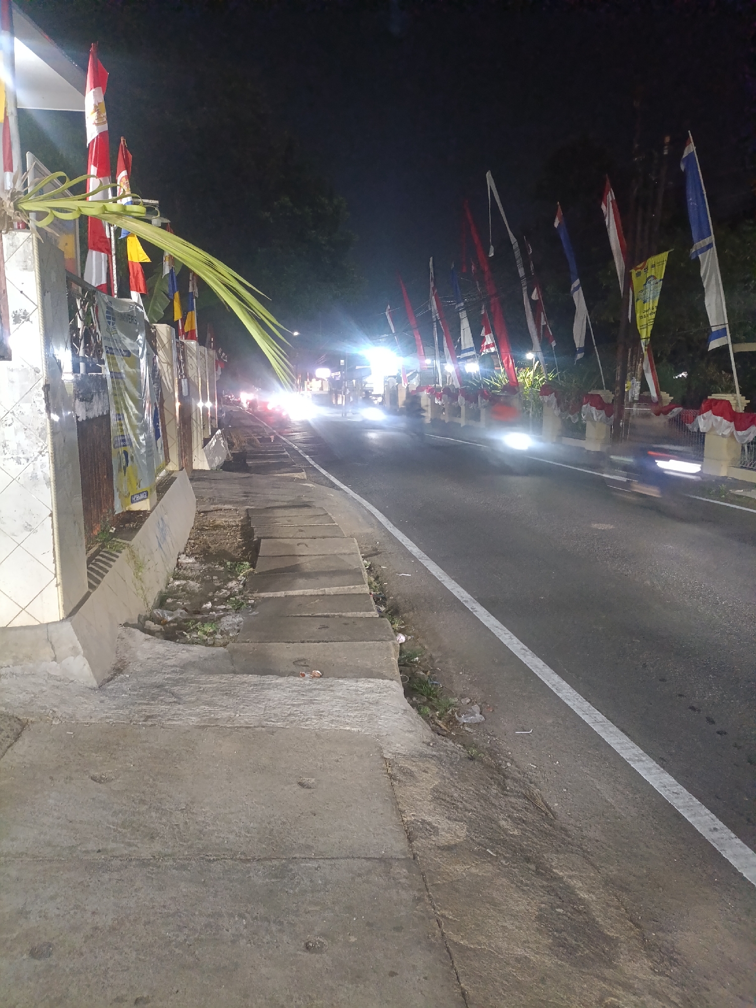 An image taken at night on a street in Jonggol, Indonesia, shows a road with traffic and sidewalks decorated with flags. The scene is illuminated by streetlights and vehicle headlights, creating a dynamic atmosphere.
On the left side of the image, a sidewalk area is visible, featuring a low concrete wall with decorative tilework. A large palm frond hangs prominently from a structure near the sidewalk. Beyond this, a series of flags are mounted, some red and white, others with different colors and designs. Banners with text are also present, though illegible due to the lighting and resolution.
The road itself is paved and marked with a white line on the right side. Several vehicles are on the road, with their headlights creating streaks of light due to the long exposure of the photograph. The movement of these vehicles, including what appears to be a motorcycle on the right, is captured as motion blur.
Along the right side of the road, a decorative railing is visible, adorned with red and white fabric draping. The area beyond the railing is obscured by darkness and trees. The overall impression is a street prepared for a celebration or event, with an emphasis on national or local pride through the display of flags and decorations. The presence of traffic indicates the street is still actively used despite the late hour.