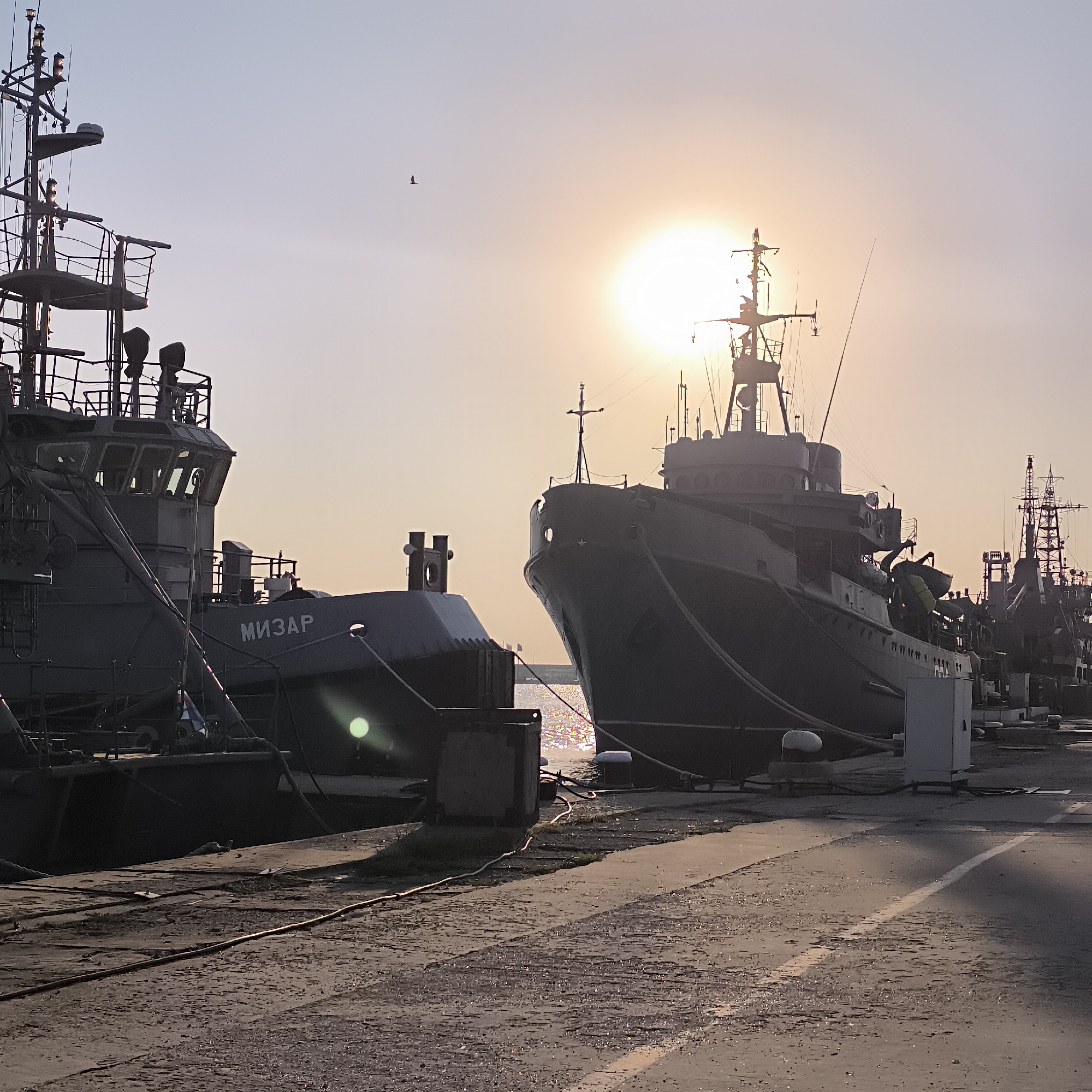 The image depicts a waterfront scene at sunset in Varna, Bulgaria. Two large ships are docked at a pier. The ship on the left has "МИЗАР" written on its hull. The sun is low in the sky, casting a warm glow and creating lens flares on the water and parts of the ships. The sky is clear with a hint of haze. There are no visible people in the image. The environment is industrial, with concrete piers and mooring lines visible. Other ships and structures are visible in the background, suggesting a port or naval base. The overall mood is tranquil, with the setting sun creating a serene atmosphere.