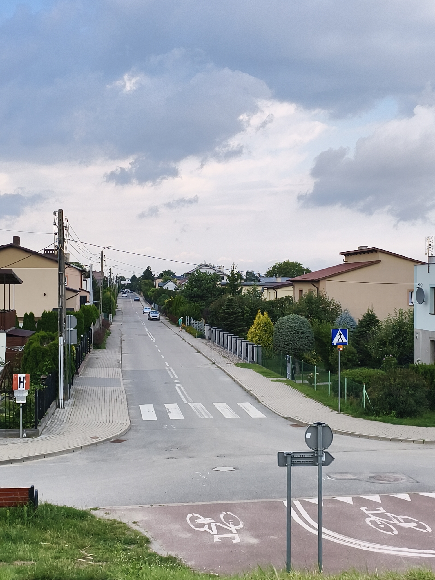 The image shows a straight, asphalt road in Tczew, Poland, lined with houses and trees. The road has white lane markings and a pedestrian crossing. Several cars are visible on the road, and a person is walking on the sidewalk. The sky is cloudy. There is a bus stop sign and a bicycle lane. The image is taken from an elevated position looking down the road.