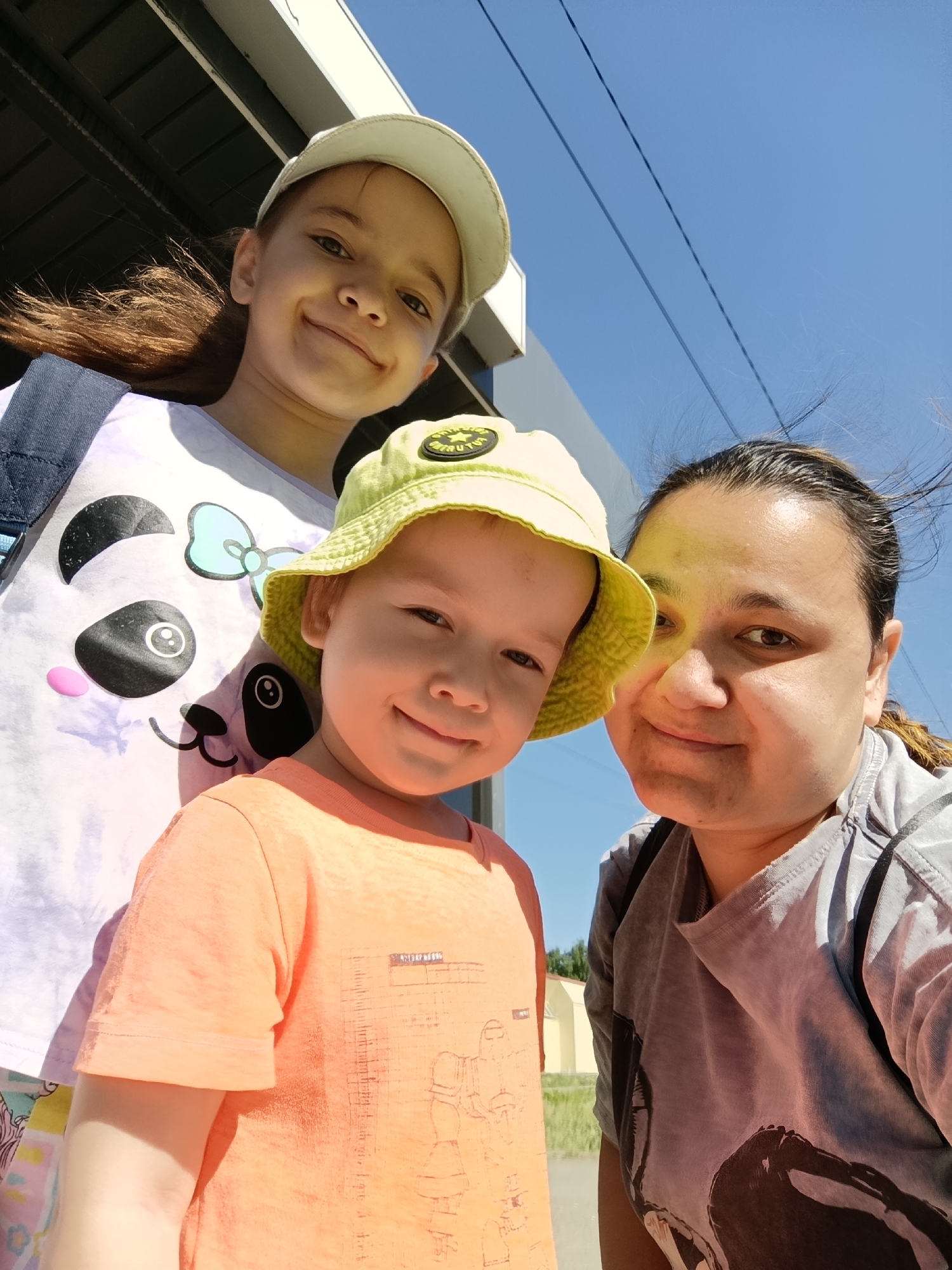 Three people are in a selfie outdoors on a sunny day. The person on the right appears to be a woman, wearing a gray shirt. A younger child is in front, wearing a bright orange shirt and a yellow hat. An older child wearing a white shirt with a panda bear graphic and a white baseball cap is to the left. They appear to be under an overpass, with blue sky visible above. The location is Chaikovsky, Russia.