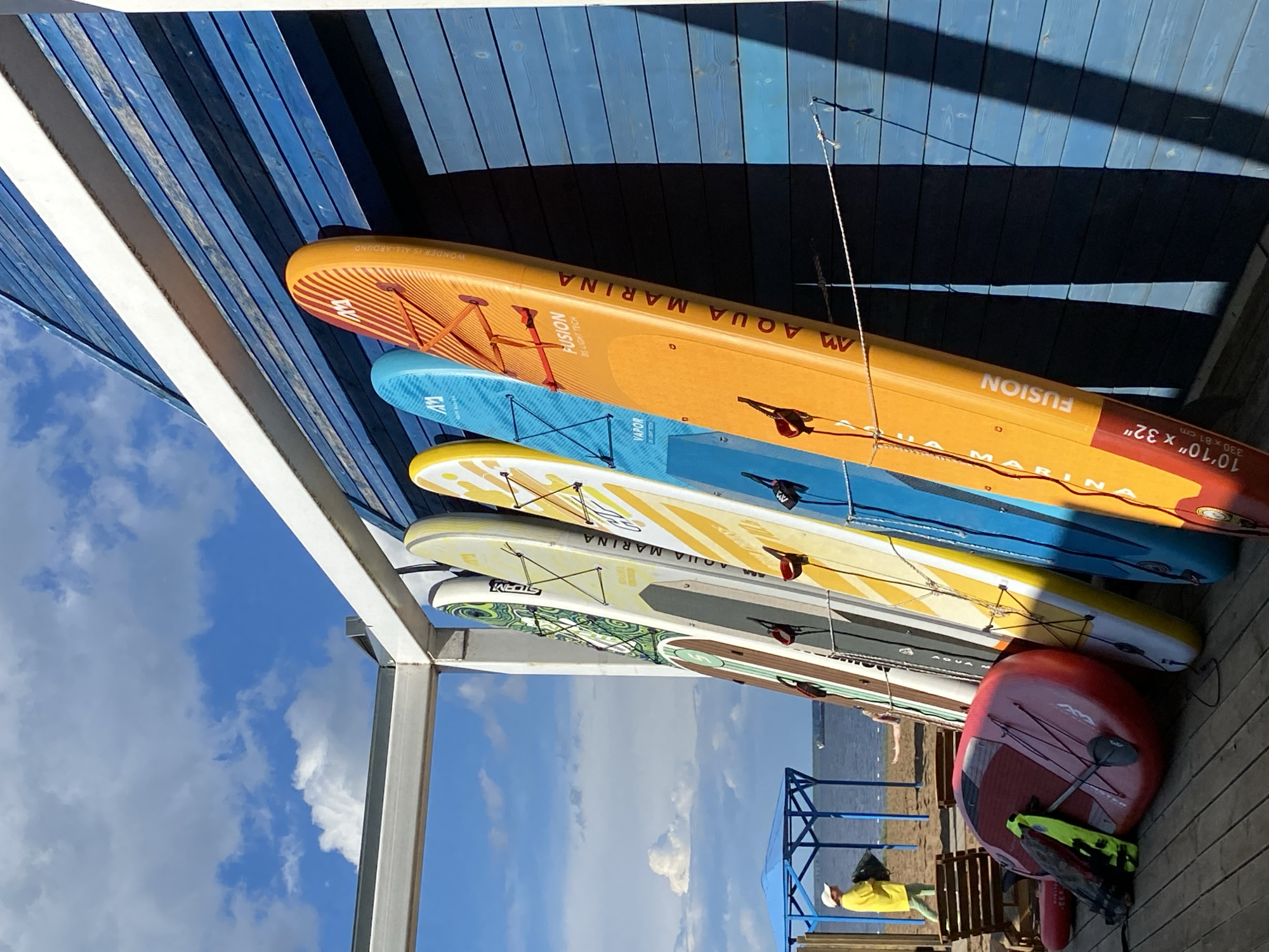This media file depicts a collection of stand-up paddleboards (SUPs) stacked and leaning against a blue wooden structure. The SUPs are of various colors and designs, including orange with red stripes, bright blue, yellow with white patterns, and white with green and brown accents. Some of the SUPs have visible text, such as "AQUA MARINA FUSION," "VAPOR," and "STORM," along with dimensions like "10'10" x 32"" and "330 x 81 cm."
In the background, a clear blue sky with scattered white clouds suggests fair weather, likely during the daytime. The scene appears to be outdoors, possibly at a beach or waterside recreational area, given the presence of wooden planks and the distant view of water.
In the lower right portion of the image, a red kayak or another SUP is visible, along with a yellow life vest or buoyancy aid. There are also hints of a wooden railing or structure and a person in a yellow shirt and white hat can be seen in the distance. The overall impression is one of readiness for water activities. The lighting and shadows suggest it is a sunny day.
The location context of Chaikovsky, Russia, implies this scene is likely from a recreational spot along a river or lake in that region. The image captures a moment of calm before or after water sports.