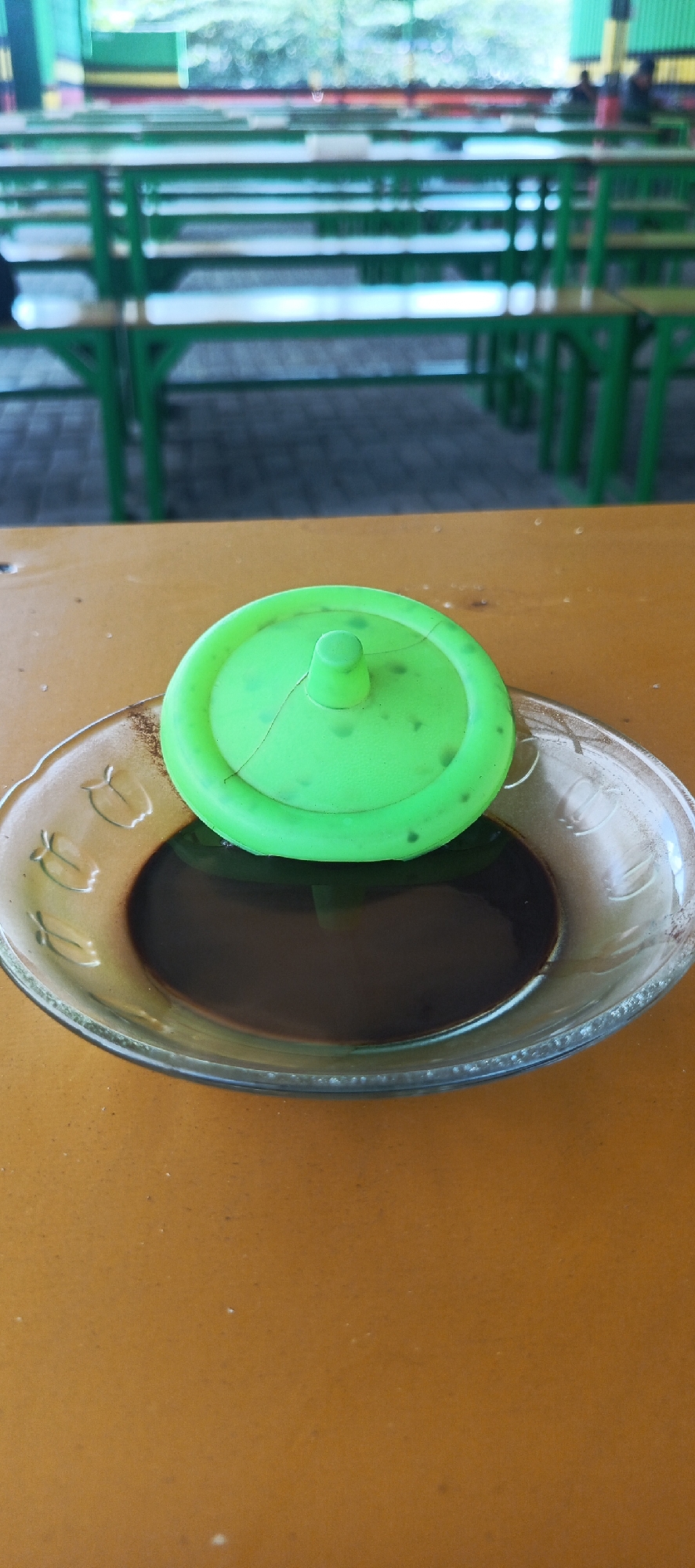 A close-up shot shows a clear glass saucer with a dark liquid, possibly coffee, on a wooden table. A cracked, bright green plastic lid is placed on top of the saucer, partially covering the liquid. In the blurred background, rows of green park benches and tables are visible, suggesting an outdoor seating area in a park or public space. The scene appears to be located in Kletek, Indonesia, as indicated by external information not present in the image itself.