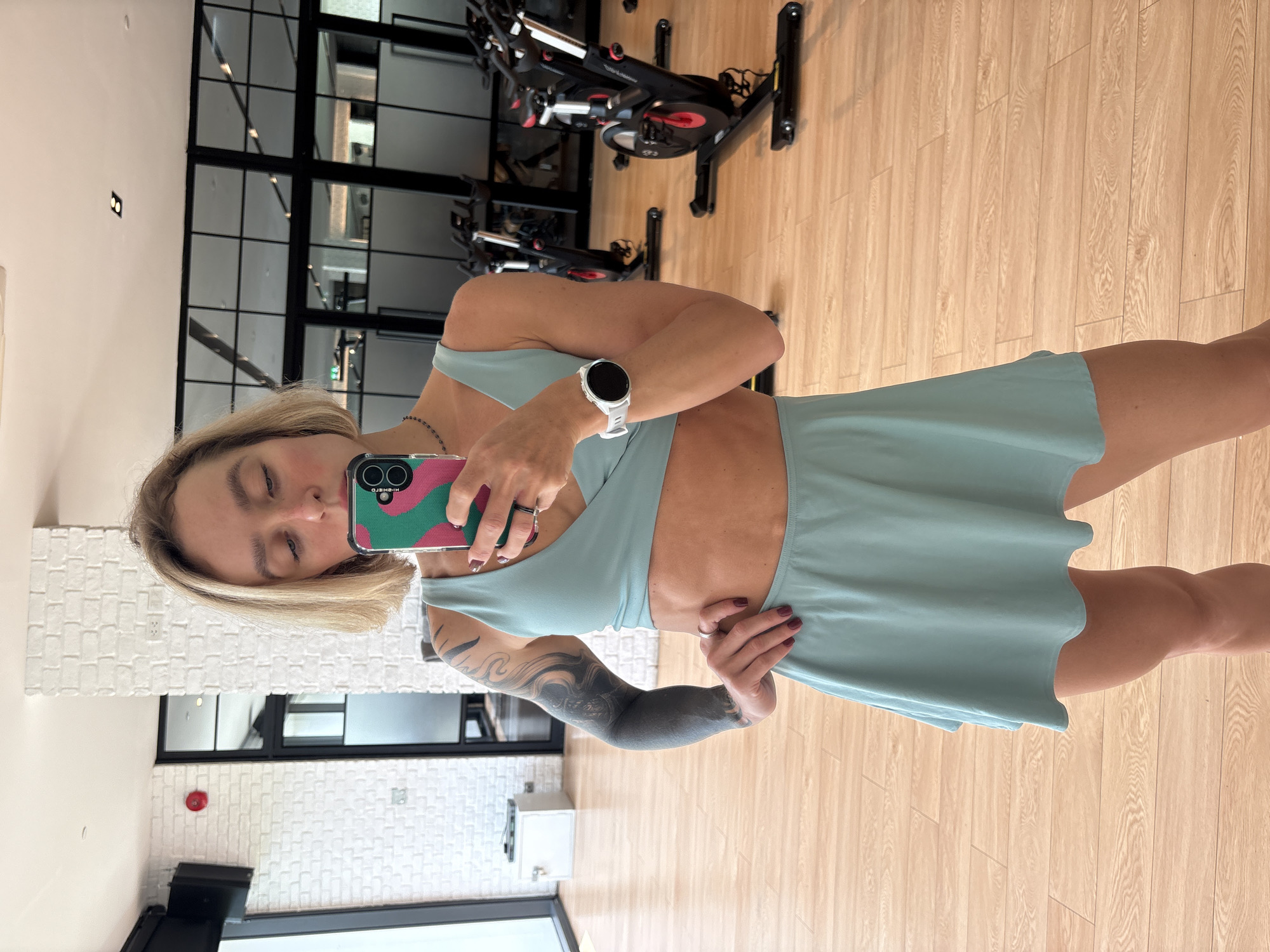 A person stands in what appears to be a gym in Rawai, Thailand, reflected in a mirror, taking a selfie. The gym features a light-colored wood floor, spinning bikes, and a wall with black-framed windows. The person is wearing a light blue athletic outfit, including a top and a skirt, and a white smartwatch. They are holding a phone with a colorful case. The person has fair skin, blonde hair, and a full sleeve tattoo on their left arm. A white brick wall and gym equipment are visible in the background through the mirror.