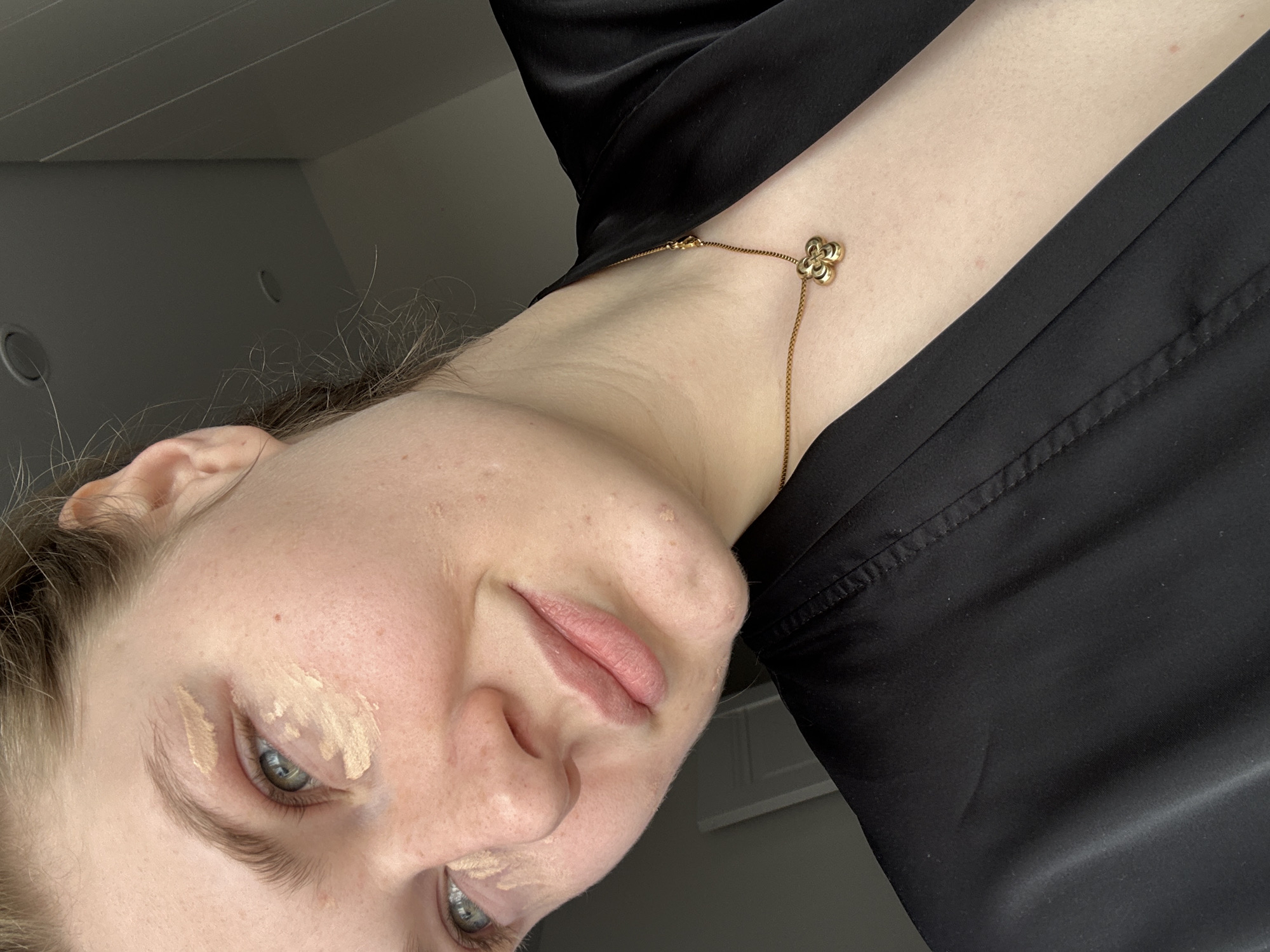 A selfie of a young woman with blonde hair, fair skin, and green eyes. The image is rotated to the left. The woman is wearing a black garment, and a gold-colored necklace with a flower pendant is visible around her neck. She has makeup applied under her eyes. The background is a neutral wall, suggesting an indoor setting. The image does not provide any details that would place it in Kazan, Russia.
