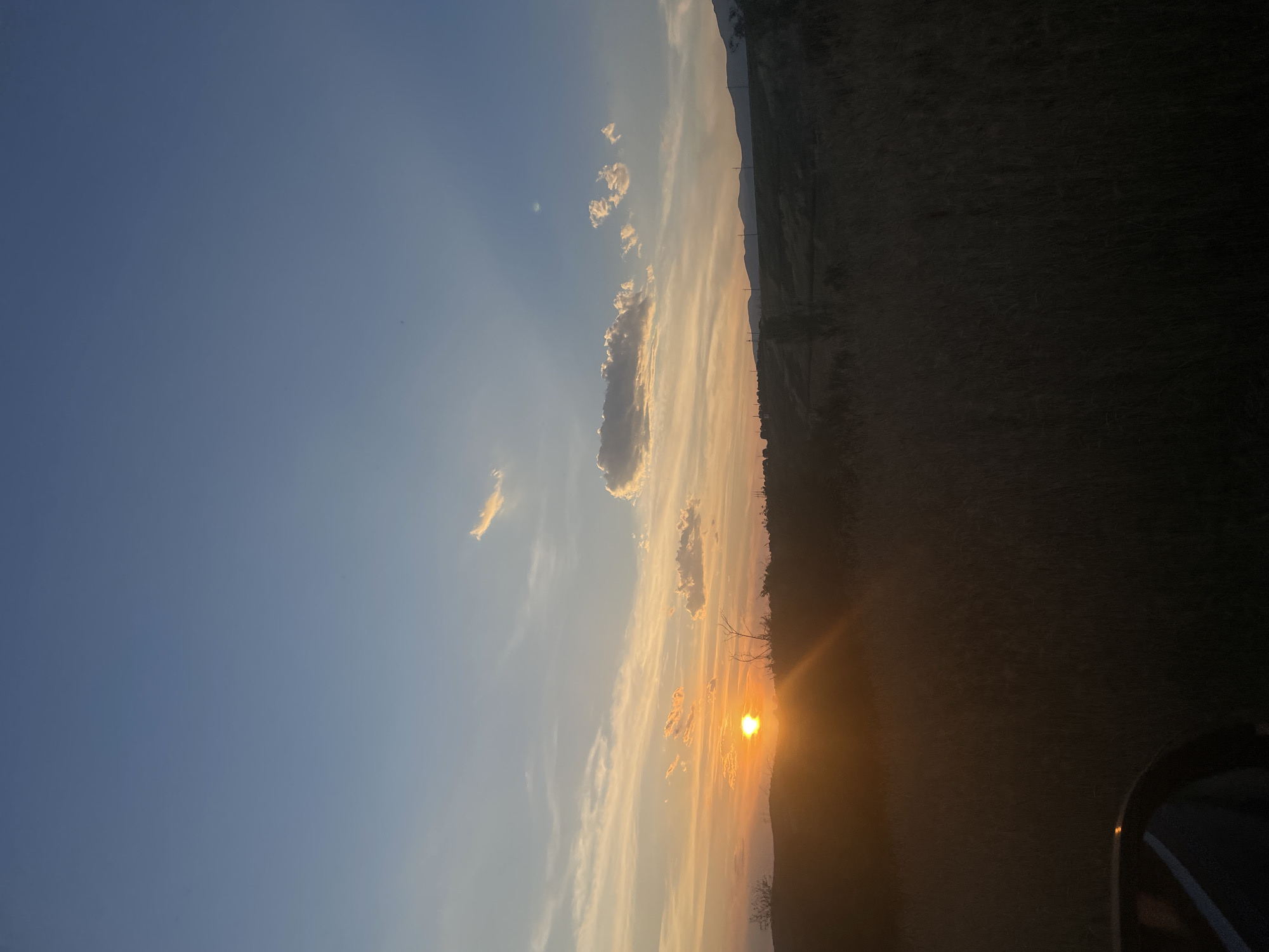 The image depicts a rural landscape at sunset, with the sun visible as a bright orb near the horizon. The sky is a gradient of blues and oranges, with scattered clouds reflecting the sunlight. A silhouette of hills or mountains can be seen in the distance. The foreground is dominated by tall grass and vegetation, casting the immediate area in shadow. A portion of a vehicle, likely a car, is visible in the lower left corner. The scene appears tranquil and natural, with no specific structures or activity indicating an urban environment. The reference to Domlyan, Bulgaria, suggests the photograph was taken in or around that location.