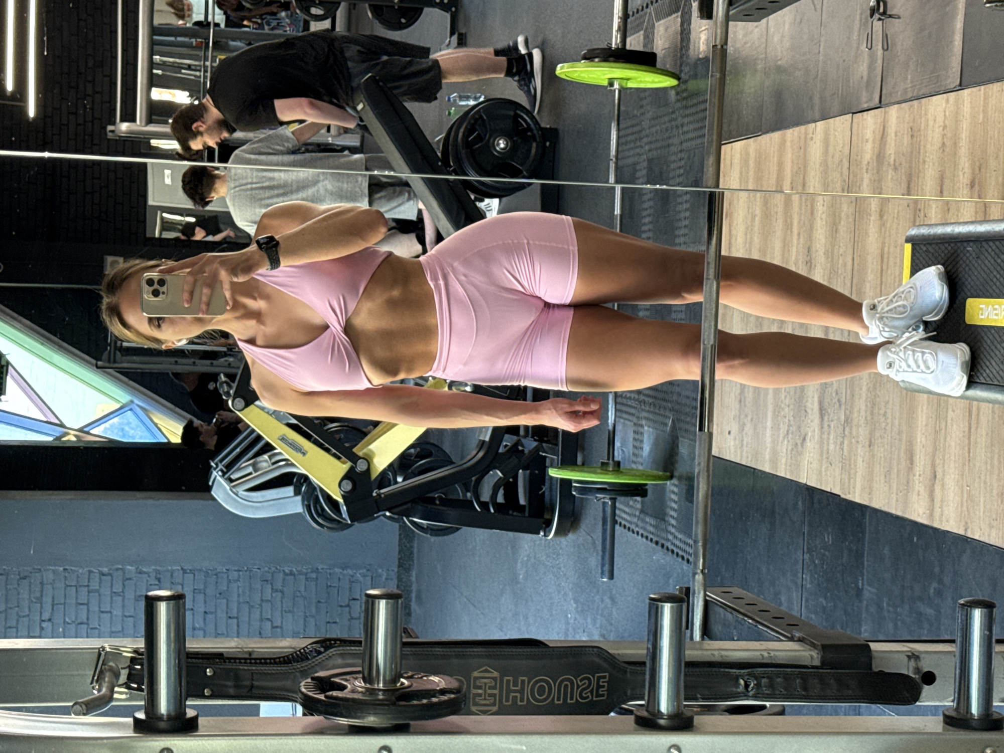 A woman is taking a mirror selfie in a gym. She is wearing a pink sports bra and pink shorts. She is standing on a yellow platform with her white sneakers. In the background, there are gym equipment, including a weight rack and barbells. There are also other people in the gym. The gym appears to be located in Saint Petersburg, Russia.