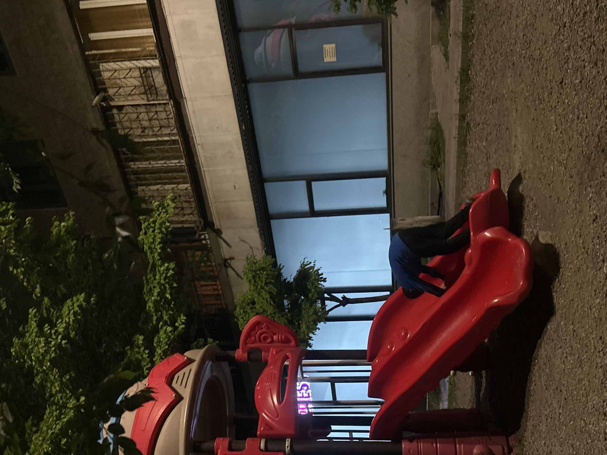The image shows a playground in Yerevan, Armenia, at night. A child wearing a blue shirt and black pants is climbing on a red slide. The playground is surrounded by trees and gravel. In the background, there is a building with windows and a balcony.