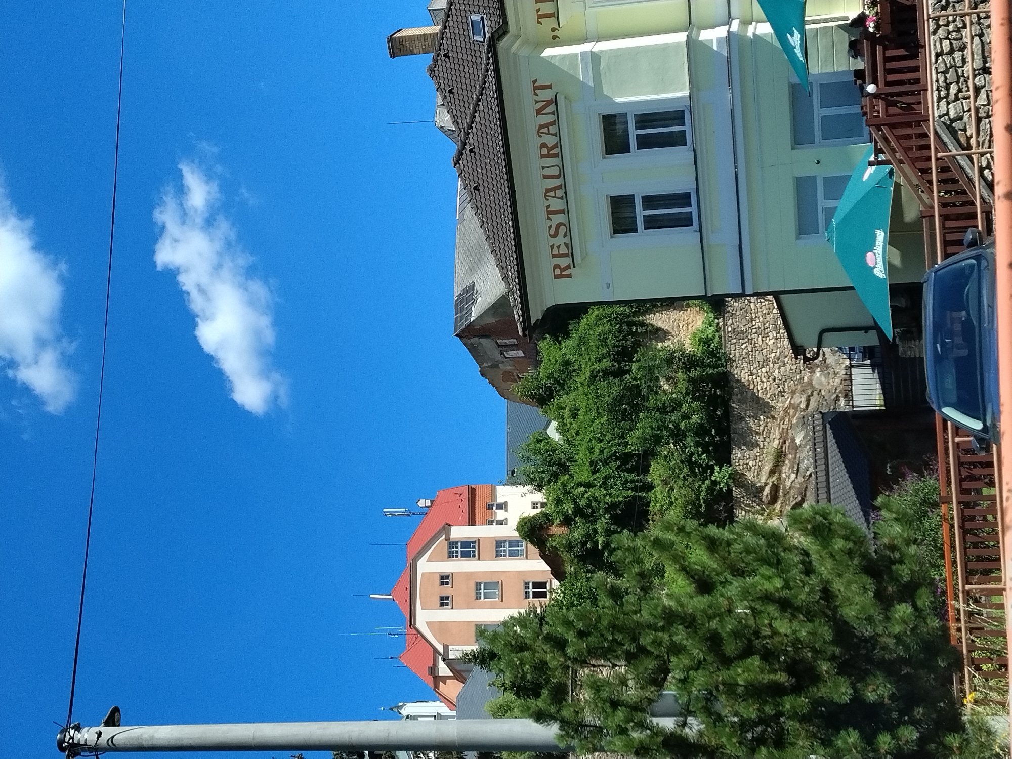 The image shows a street scene in Vimperk, Czechia, on a sunny day. In the foreground, a dark-colored car is partially visible, parked behind a wooden fence. A bright green umbrella with the Pilsner Urquell logo is open next to a restaurant building. The building, painted a light yellow, has "RESTAURANT" written in large brown letters above its windows. Behind the restaurant, a stone wall supports a hillside covered in lush green trees and vegetation. Further back, a multi-story building with a red roof is visible, and to its right, another building with a dark tiled roof. The sky is clear blue with a few white clouds. A tall concrete utility pole with wires stretches across the upper left portion of the frame.