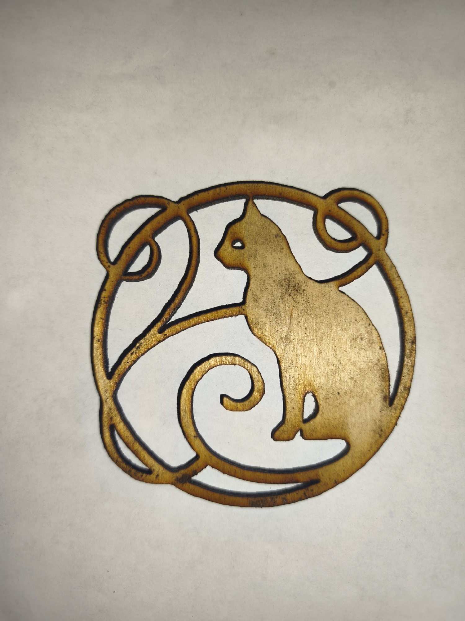 A close-up, overhead shot shows a golden-brown, laser-cut wooden ornament featuring a silhouette of a cat within an art nouveau-style circular frame. The cat is sitting and facing left, with its tail curled around its body. The frame consists of flowing, organic lines with small circular embellishments. The ornament has a slightly distressed appearance with visible wood grain and dark edges from the laser cutting process. The background is a plain, off-white surface with subtle texture and a few faint, dark speckles. No specific location in Гаранскі сельскі Савет, Belarus can be identified from this image.