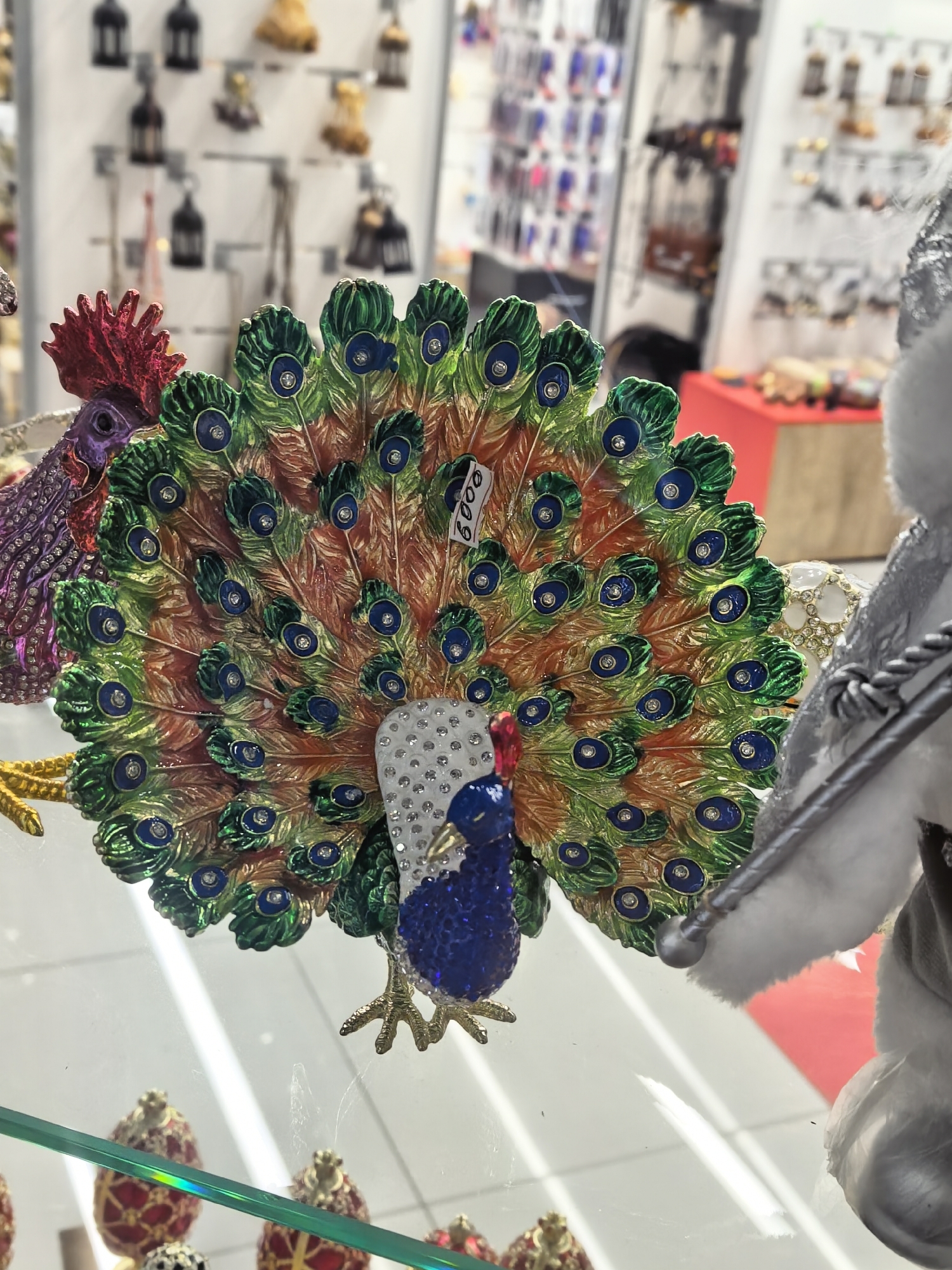 A close-up shot displays a decorative peacock figurine with an elaborate tail, showcasing green, orange, and brown feather patterns punctuated with blue and silver gem-like accents. The peacock has a blue body and a white, jeweled chest. A price tag reading "6000" is attached to the tail. The figurine stands on a clear glass shelf, with a few decorative eggs visible below. In the blurry background, other decorative items are displayed on shelves, suggesting a retail environment. Red and white items appear in the background, possibly holiday decorations. The items are in a store location in Moscow, Russia.