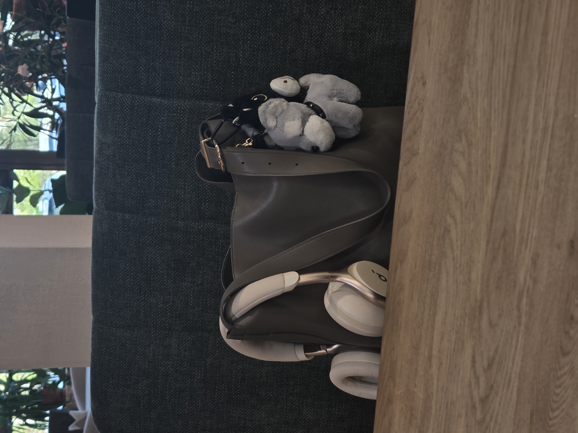 The image, rotated 90 degrees to an upright position, presents a close-up view of personal belongings resting on a dark green, textured fabric surface, likely a sofa or armchair.
Centrally, a dark grey or olive-green shoulder bag is visible, with its strap and a gold-colored buckle prominent on the left side. Nestled within and around the bag's opening are several small, plush keychains or charms, including two grey ones, a black one, and a white one with simple facial features.
To the right of the bag, a pair of light-colored (beige or off-white) over-ear headphones with gold accents is placed, partially tucked into the bag. A distinctive lowercase 'b' logo, associated with Beats by Dr. Dre, is visible on the outer shell of one earcup. A light brown wooden surface, possibly a table or desk, frames the right edge of the composition.
In the background, beyond the dark green fabric, a window allows natural light into the scene, with green foliage from a plant visible outside, suggesting an indoor setting during the daytime. The overall impression is that of everyday items casually placed aside in a living or lounge area. No individuals or specific activities are depicted.
