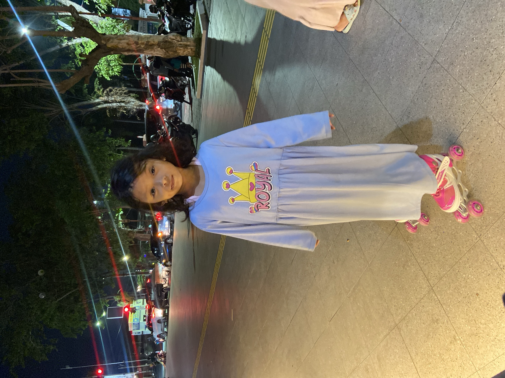 A young girl wearing a light blue dress with a crown and the word "ROYAL" printed on it stands on roller skates on a sidewalk at night. She is facing the camera with her hands at her sides. The street behind her is lit by streetlights and the headlights of cars. Several motorcycles are parked on the side of the road. The scene is outdoors in a city environment. The location is identified as Sidoarjo, Indonesia.