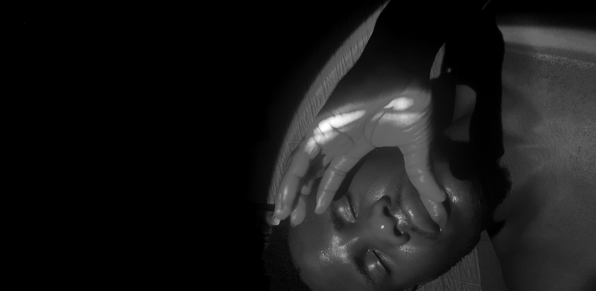 The image features a close-up, monochromatic portrait of a person, likely an adult, taken in a low-light setting in Narayi, Nigeria. The person's head and neck are visible, positioned at an angle, with a hand resting over their eyes and forehead, partially obscuring the upper face. The eyes appear closed or are fully covered by the hand.
Dramatic, high-contrast lighting illuminates the scene, with a strong, narrow beam of light highlighting the palm and fingers of the hand, as well as parts of the forehead, nose, and lips. This light creates stark shadows across the face and on the textured surface behind the person, which appears to be a wall or floor. The skin on the face and hand shows a reflective sheen, possibly indicating sweat or moisture. Much of the image is enveloped in deep black shadows, particularly on the left side, adding to the somber and intimate atmosphere.
The activity depicted suggests a moment of rest, sleep, or contemplation. The hand covering the eyes could imply a desire for privacy, exhaustion, or shielding from light. No objects other than the person and their hand are clearly identifiable, and there is no visible text. The overall mood conveyed by the composition and lighting is one of quiet introspection or vulnerability.