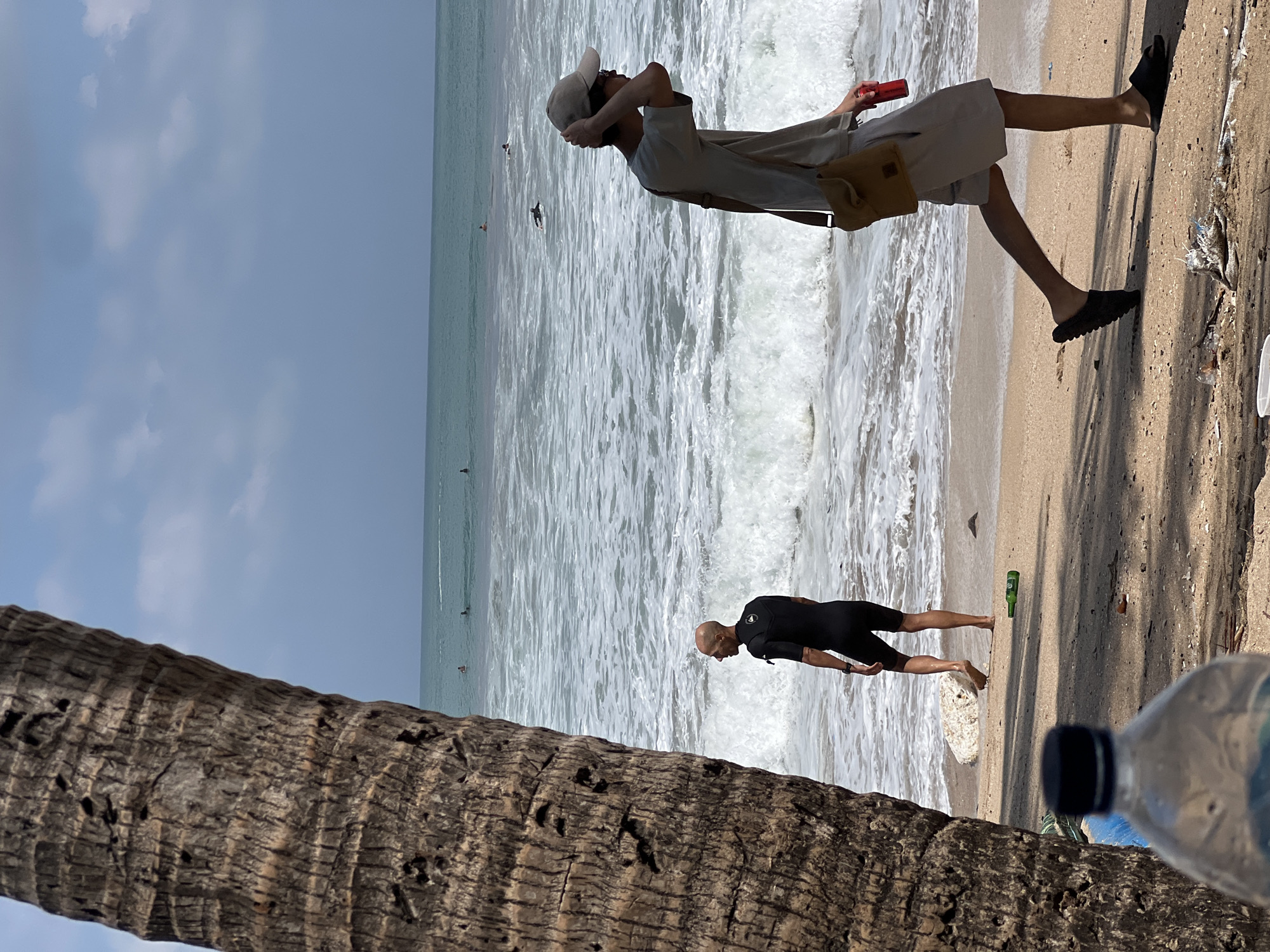 The image captures a sunny day at a beach in Kuta, Indonesia. In the foreground, the textured trunk of a palm tree partially obscures the view. To the right, a person walks along the sandy shore, wearing a light-colored t-shirt, shorts, a cap, and sandals. They carry a cross-body bag and a red can. In the middle ground, another person stands near the water's edge, wearing a black wetsuit. The ocean exhibits moderate waves and several surfers are visible in the distance. The beach is littered with a few pieces of trash, including a green bottle and some plastic debris. The sky is a clear blue with scattered white clouds.