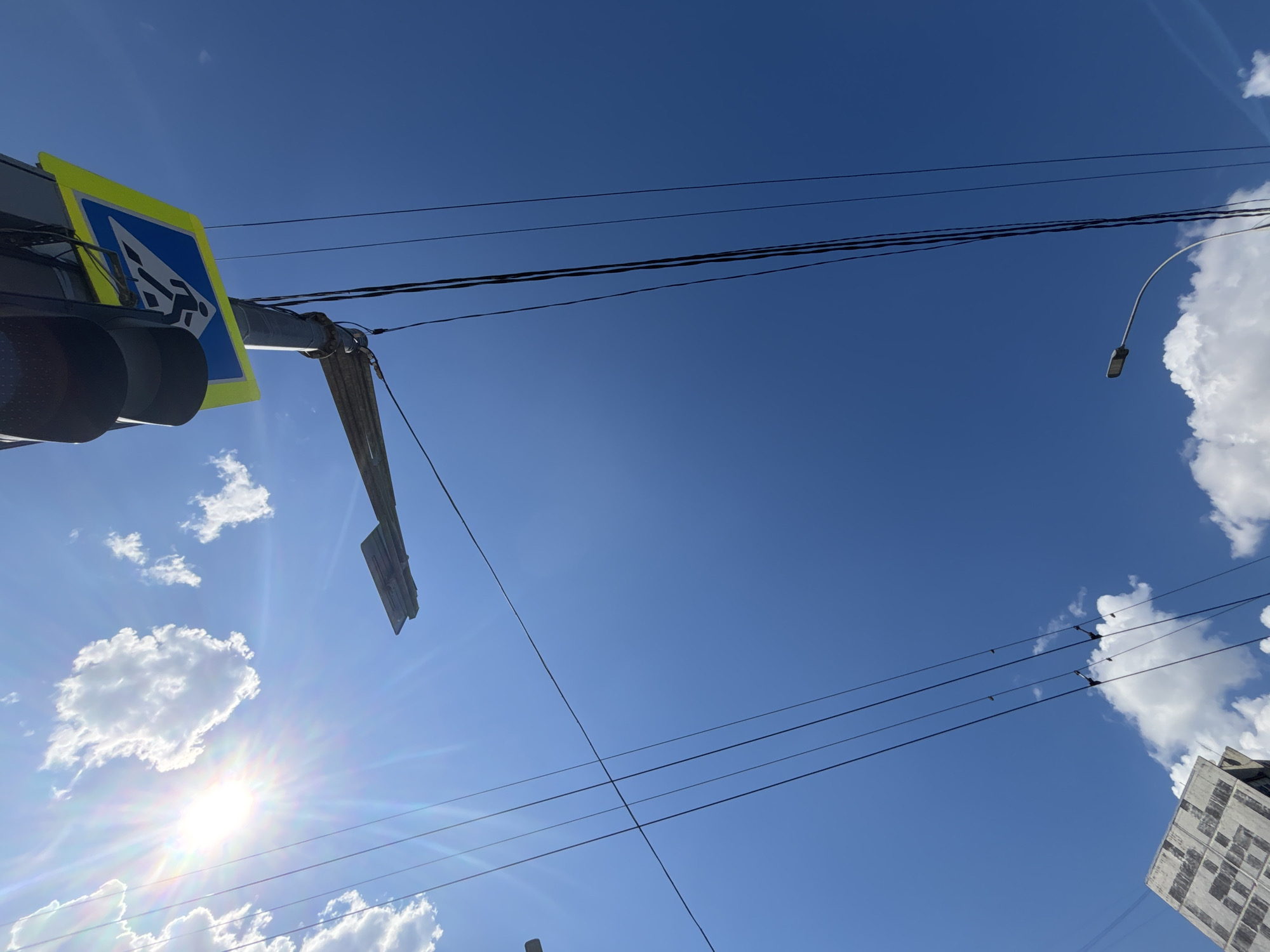 This media file depicts an outdoor urban scene on a bright, sunny day in Krasnodar, Russia. The primary focus is on a street sign indicating a pedestrian crossing, mounted on a pole alongside traffic lights. The sign is yellow and blue with a white silhouette of a person crossing the street. Overhead, a network of power lines stretches across the bright blue sky, crisscrossing and supported by poles. A street lamp is visible in the upper right quadrant. Fluffy white clouds are scattered across the sky, and the sun is shining intensely, causing lens flare and casting bright beams. In the lower right corner, a portion of a modern building with a patterned facade is visible. The overall impression is that of a typical city street with essential infrastructure, bathed in warm sunlight, suggesting it is daytime. There is no discernible activity or event taking place, and no text is legible.