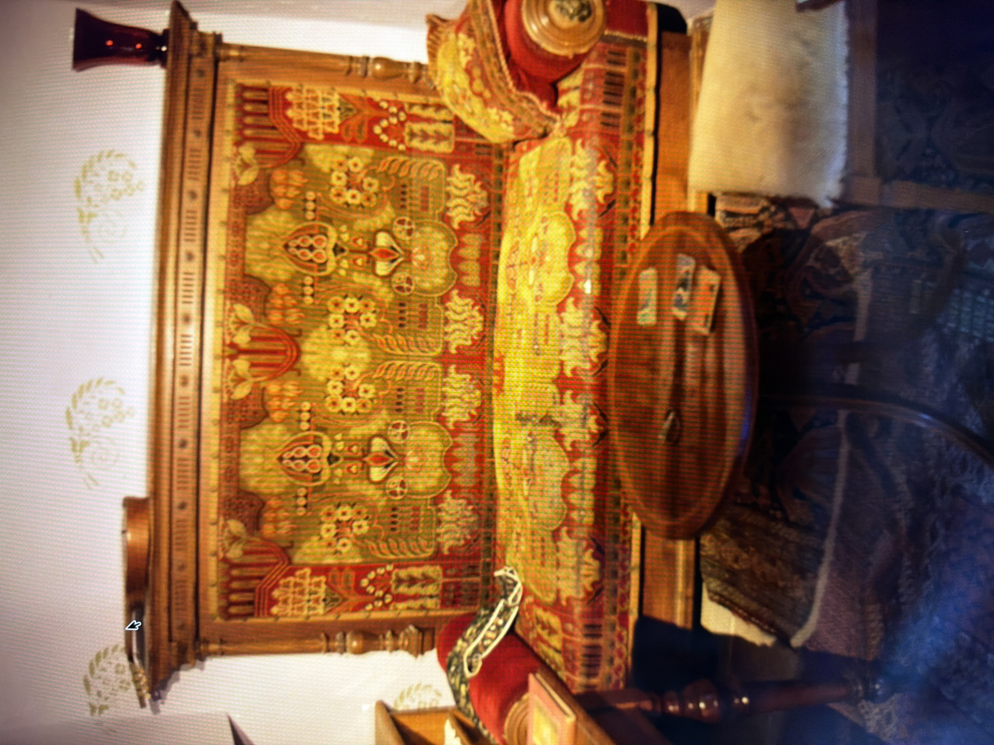 The image depicts an indoor scene, likely a room in a museum or a historical building. The central focus is an ornate, wooden-framed sofa with elaborate tapestry upholstery. The tapestry features a complex pattern of floral motifs and geometric designs in vibrant shades of red, yellow, and green. The sofa is accompanied by decorative pillows, one of which is red and the other a lighter shade with a textured surface.
In front of the sofa, a round, wooden coffee table is visible, adorned with several small, rectangular objects, possibly books or cards. The floor is covered with a patterned rug. To the left of the sofa, a portion of a wooden table or cabinet is visible, with a red book resting on it. The background shows a wall with subtle decorative stencils and a dark red vase on a shelf or mantelpiece. The lighting suggests an indoor setting.
No specific location in Kyiv, Ukraine is identifiable from the visual cues in the image. The scene is static, with no discernible actions or interactions taking place.
