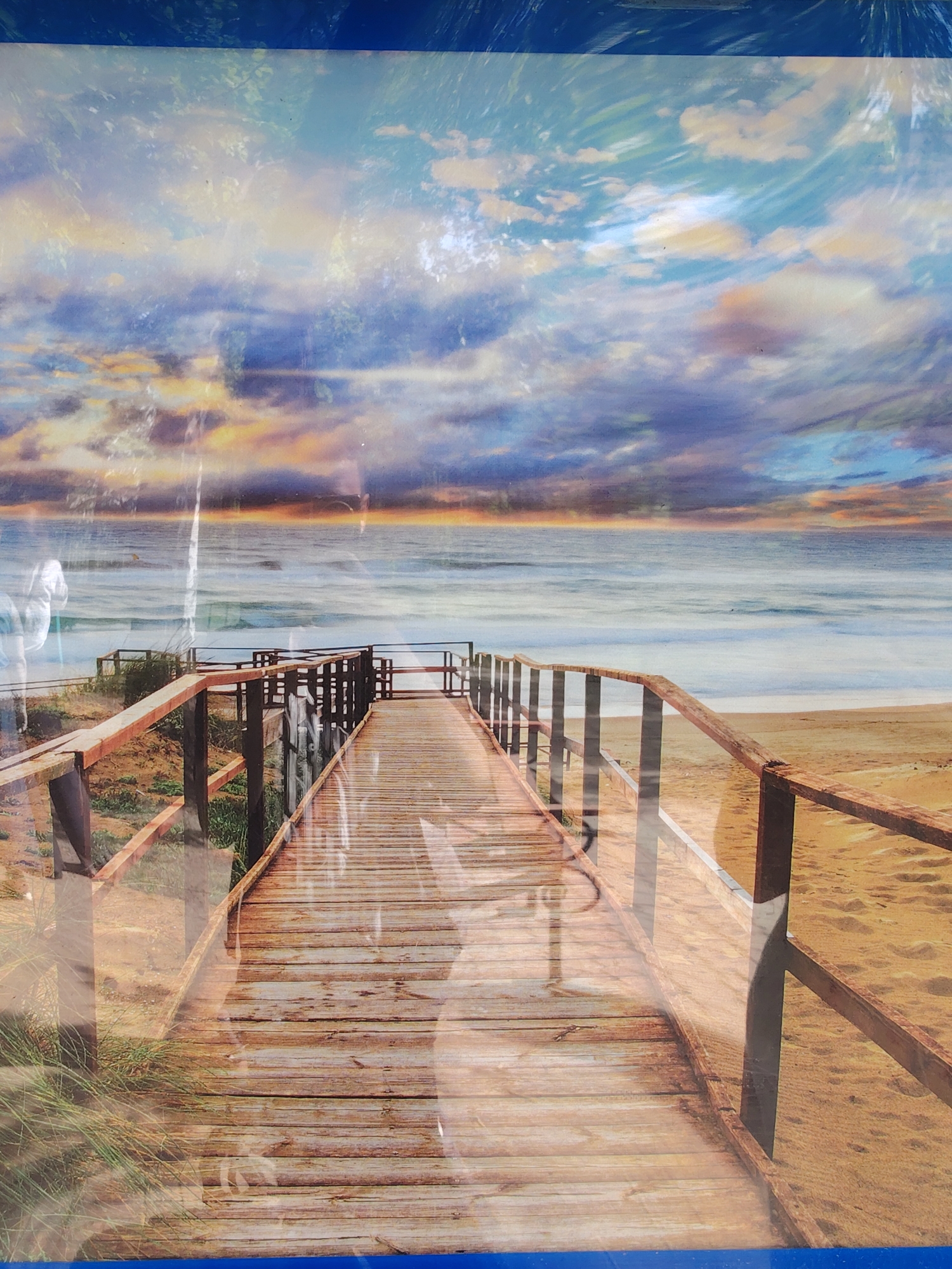 The image depicts a wooden boardwalk leading towards the ocean. The boardwalk has railings on both sides and is situated on a sandy beach. The sky is filled with clouds, with a sunset glow on the horizon. The ocean stretches out to meet the sky. It is not possible to verify the location in the city of Chaikovsky, Russia based on this image.