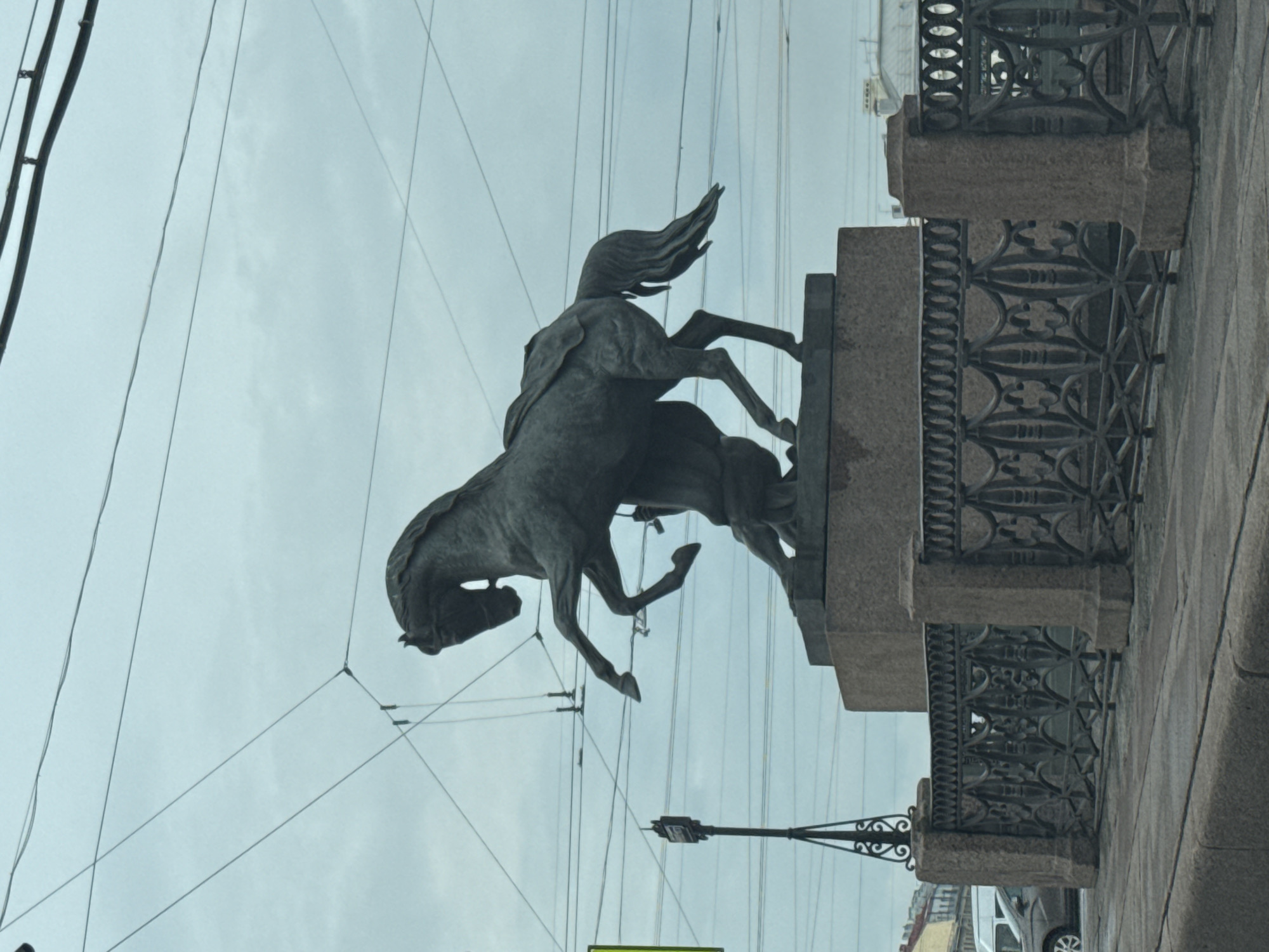 The image captures one of the horse sculptures adorning the Anichkov Bridge in Saint Petersburg, Russia. The bronze sculpture depicts a rearing horse being restrained by a man. The sculpture is mounted on a granite pedestal that forms part of the bridge's architecture, which includes a decorative iron railing. Overhead, power lines intersect the sky. A portion of the street and a parked car are visible at the bottom right of the image. The sky is overcast.